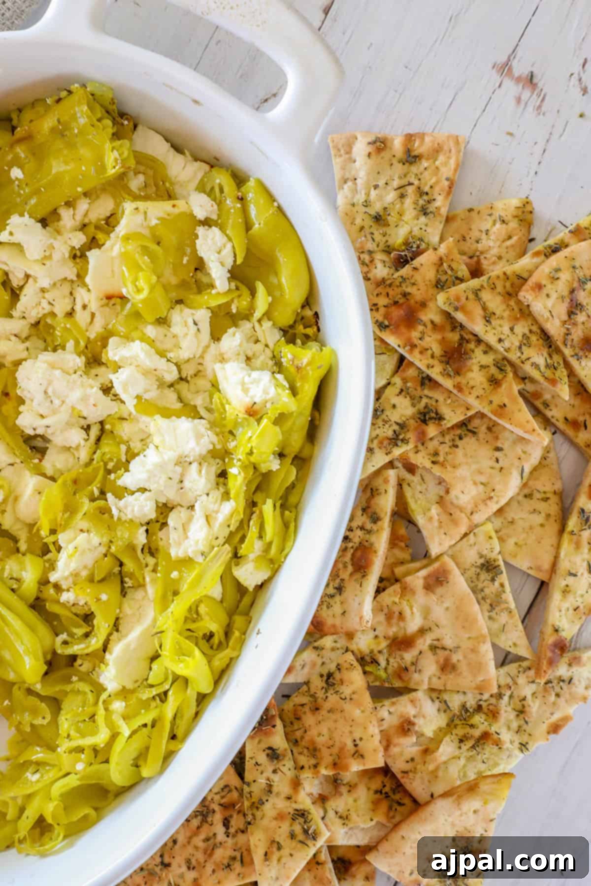 White dish with sliced pepperoncini and crumbled feta cheese, reminiscent of a pepperoncini baked feta dip, accompanied by herb-coated pita chips on a wooden surface.