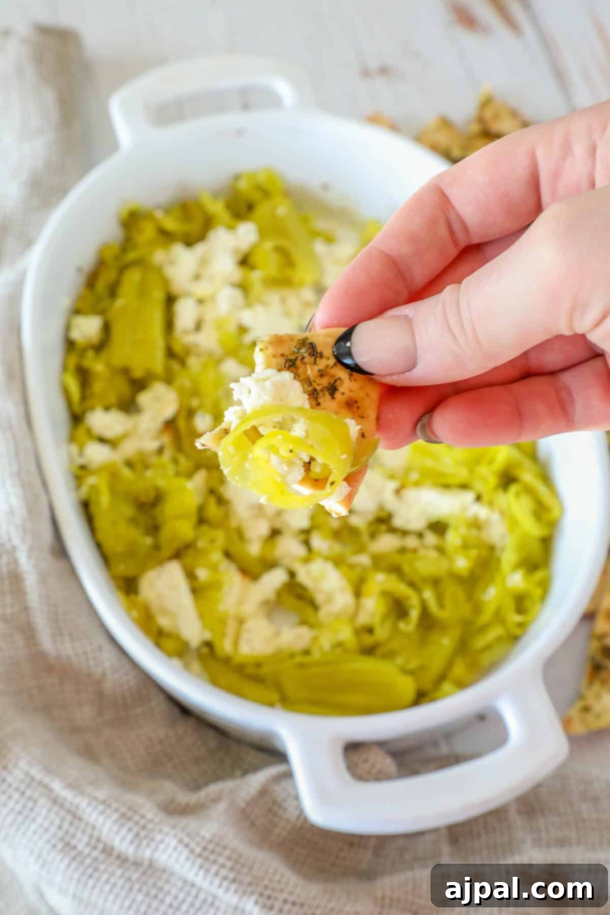A hand delicately holds a cracker crowned with green peppers and feta cheese, hovering above a white dish brimming with the delightful ingredients of a pepperoncini baked feta dip.