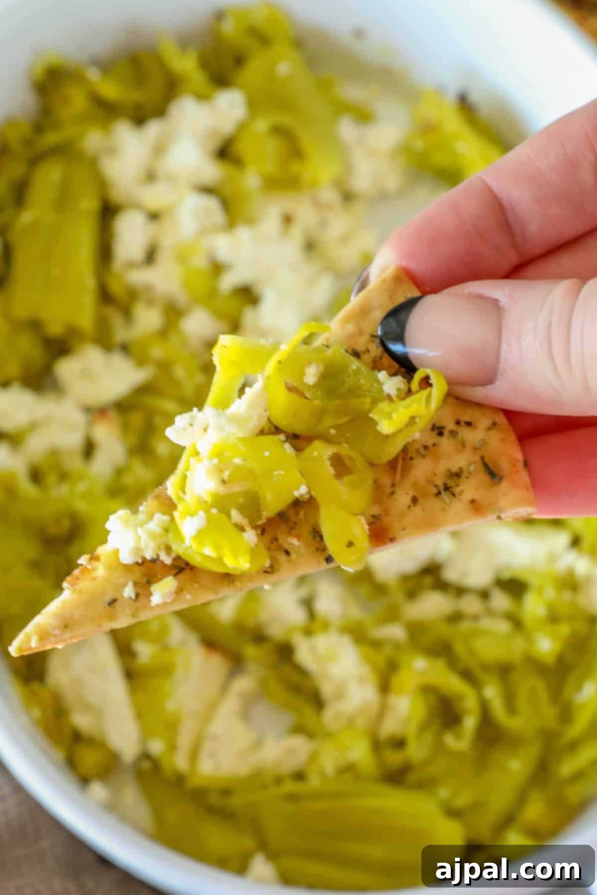 A hand holds a chip topped with sliced green peppers and crumbled cheese over a bowl of pepperoncini baked feta dip.
