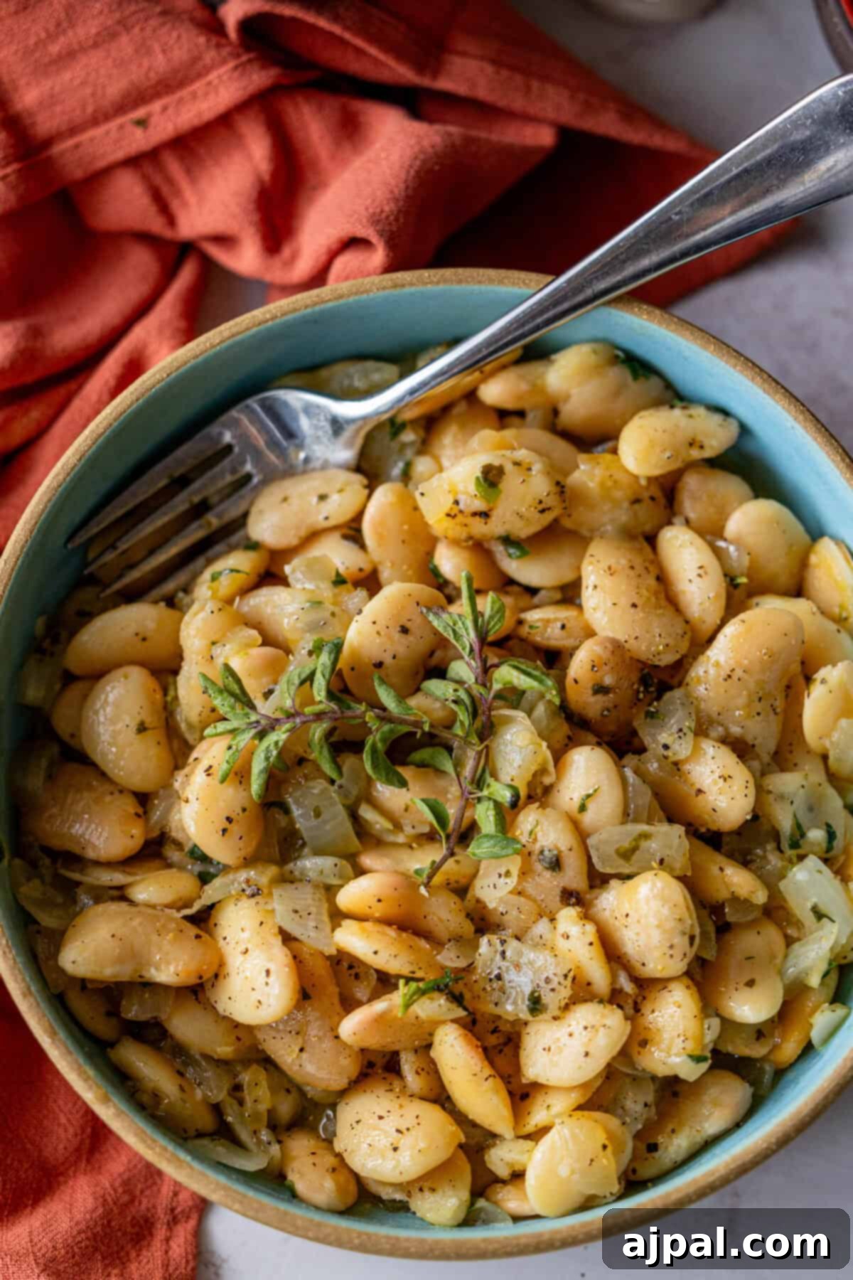 A bowl of cooked butter beans garnished with oregano and herbs, served with a fork on the side. An orange cloth is partially visible in the background.