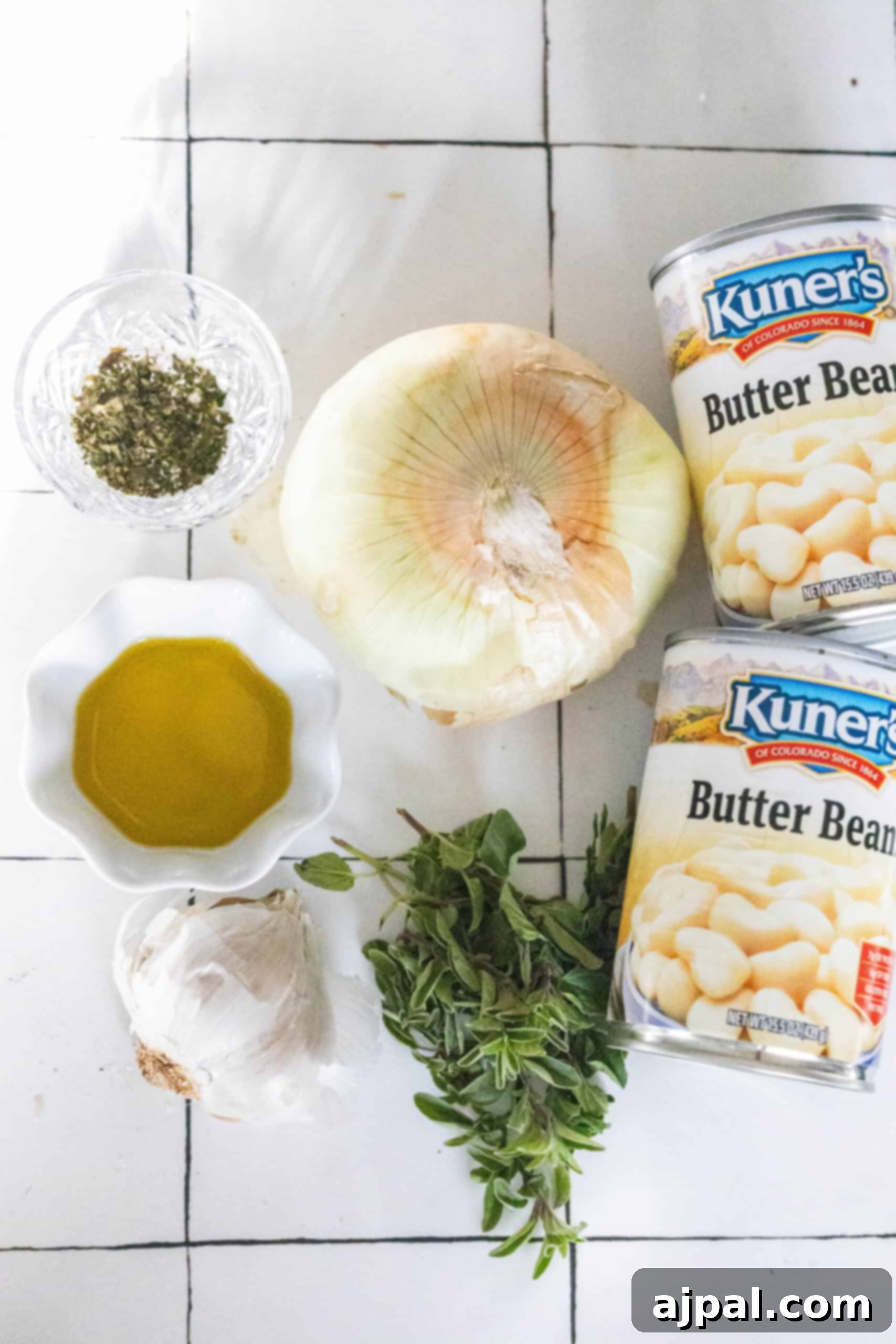 Cans of butter beans, an onion, garlic, and olive oil sit with dried oregano in a glass dish, accompanied by fresh green herbs on a tiled surface.