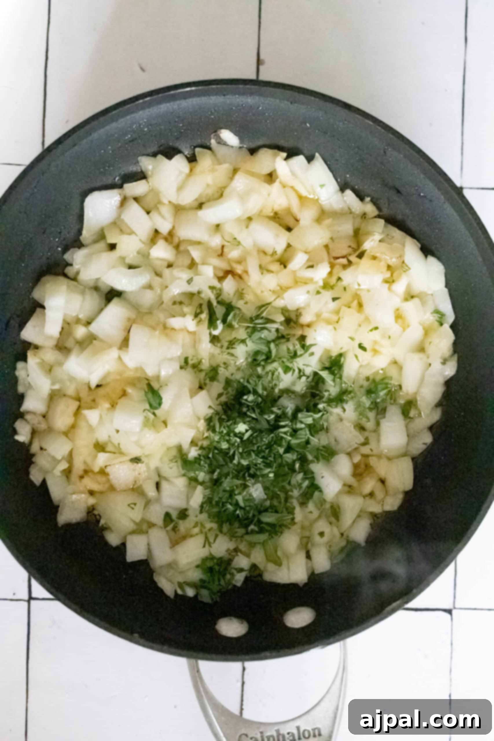 A frying pan with chopped onions, garlic, and herbs sits on a white tiled surface.