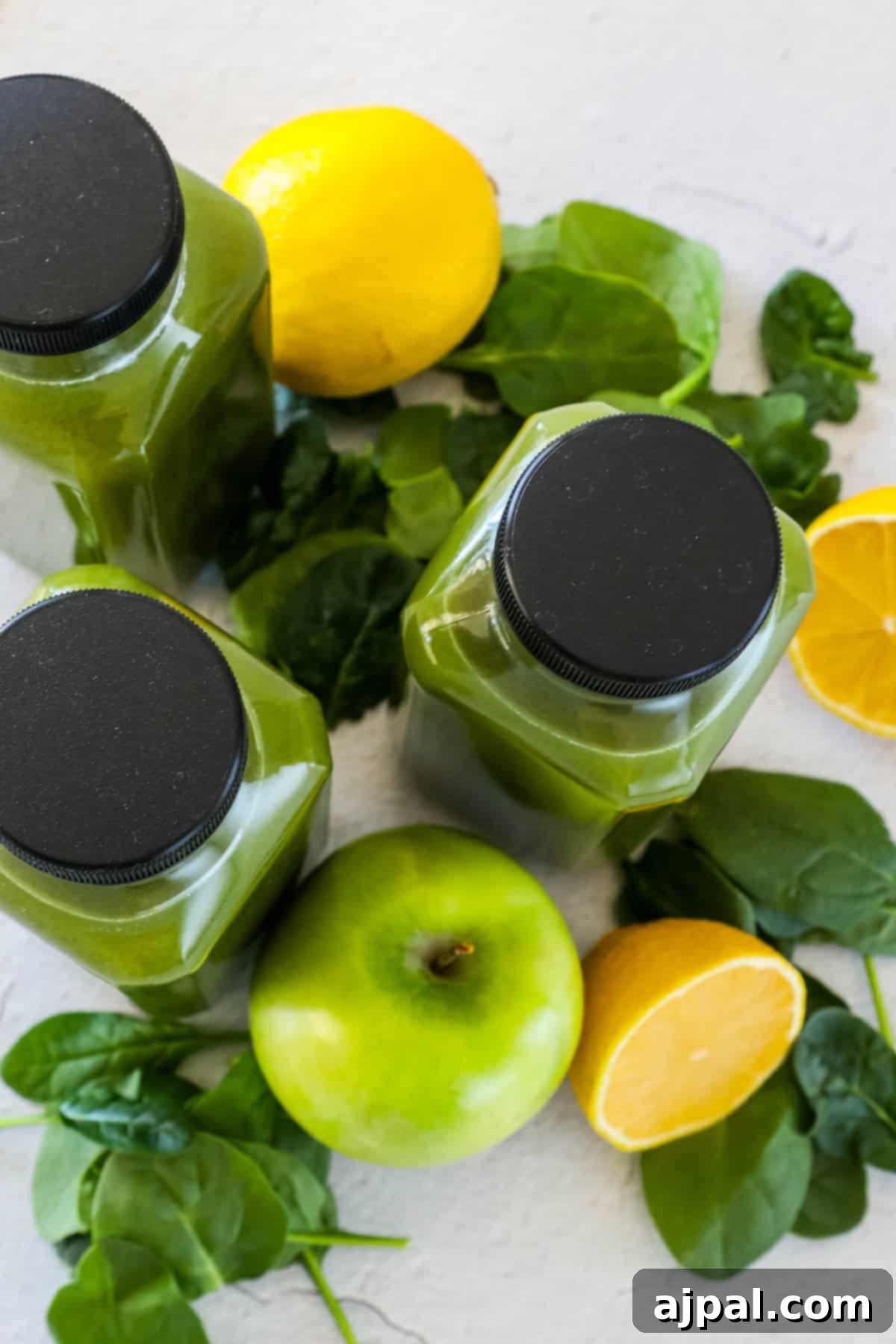 Green Shield Elixir 3 Three slender bottles of vibrant green juice, surrounded by fresh spinach leaves, a bright green apple, and whole and half lemons, resting on a clean, light surface.