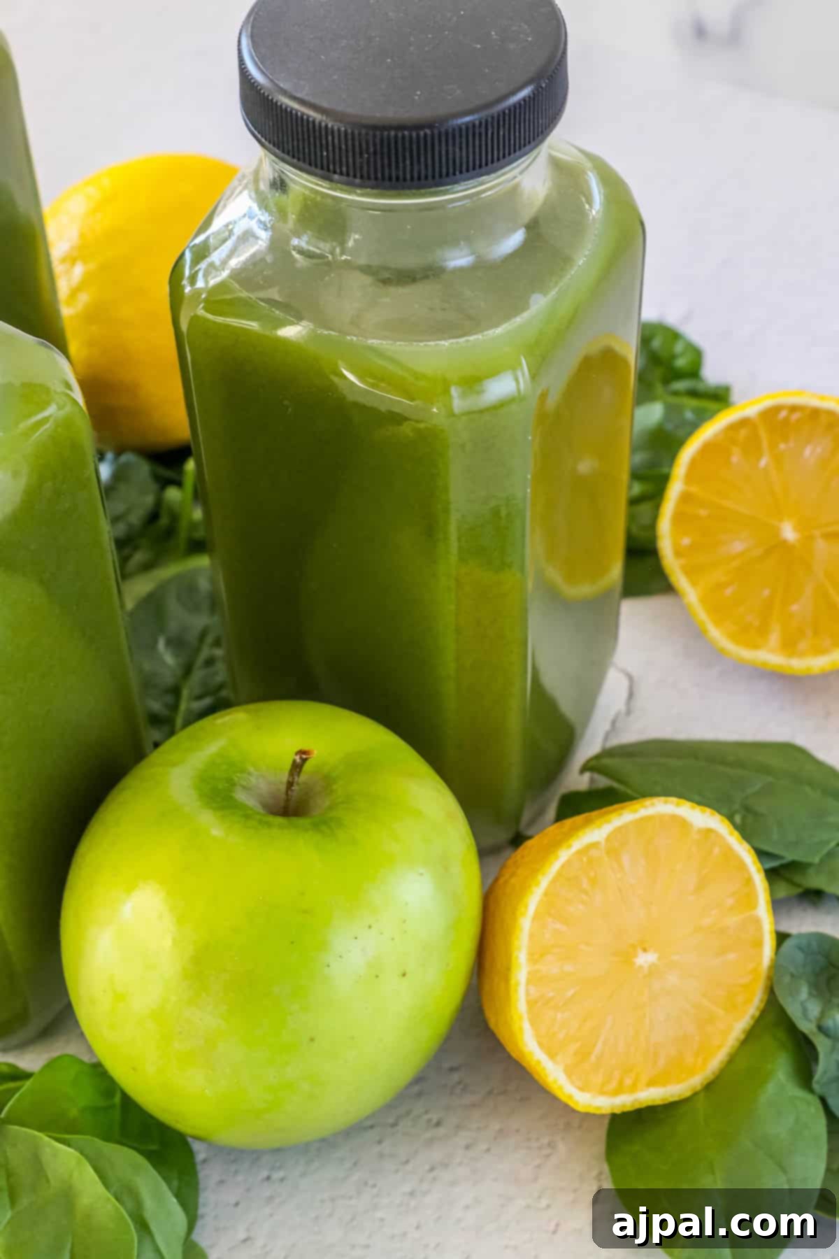 Green Shield Elixir 4 Two bottles of radiant green juice stand on a pristine white surface, flanked by a vibrant green apple, perfectly halved lemons, and fresh spinach leaves, showcasing the natural ingredients.