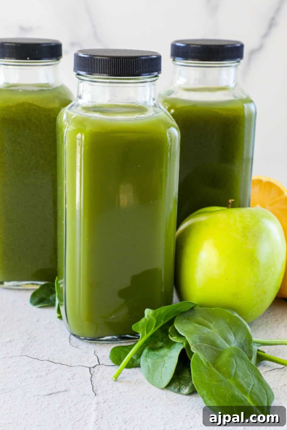 Green Shield Elixir 5 Three glass bottles filled with vibrant green juice, accompanied by a fresh green apple, half a lemon, and scattered spinach leaves, all resting on a rustic, cracked stone surface.