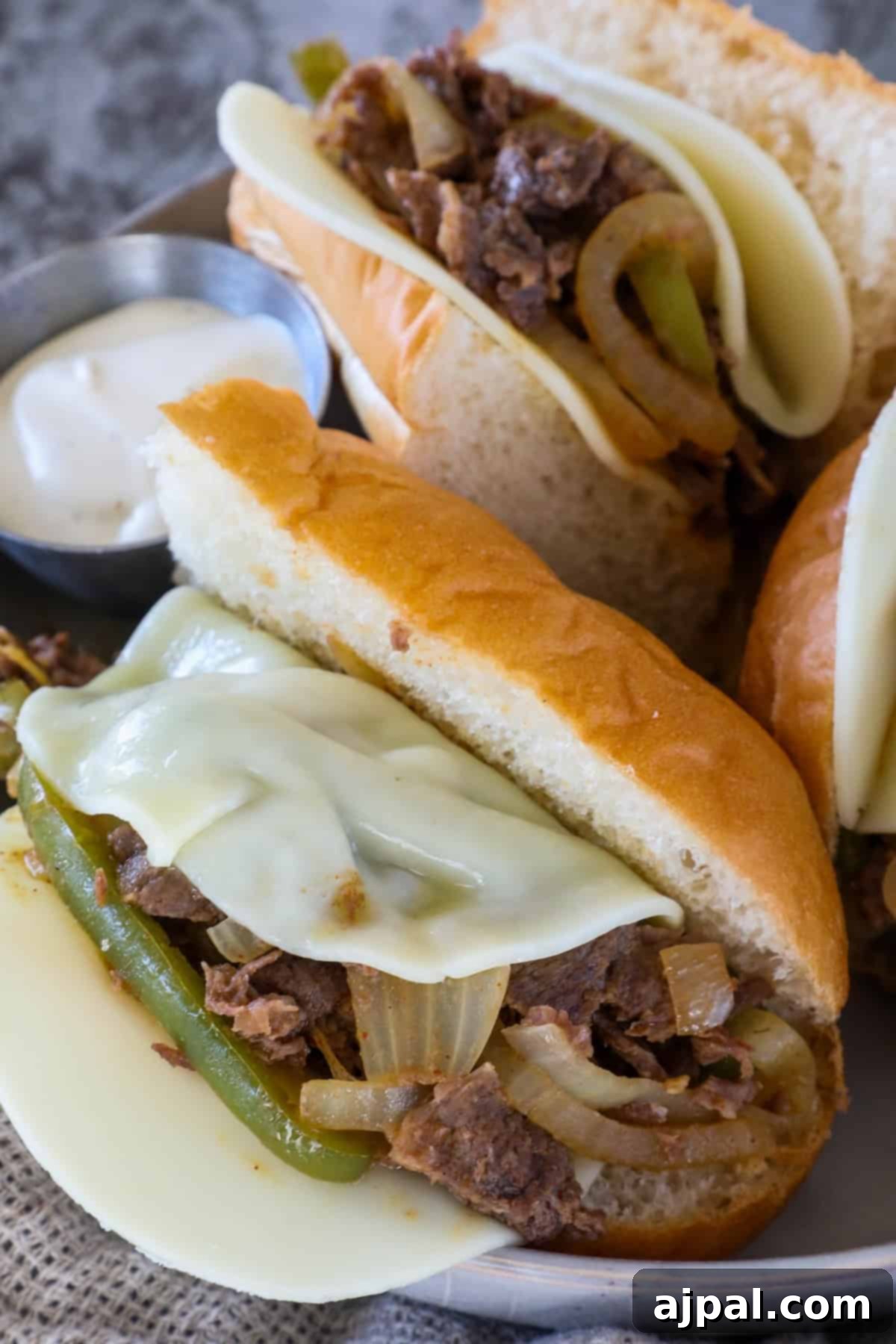 Close-up of two cheesesteak sandwiches with sliced beef, melted cheese, and sautéed onions and peppers, on a plate next to a small dish of white sauce.