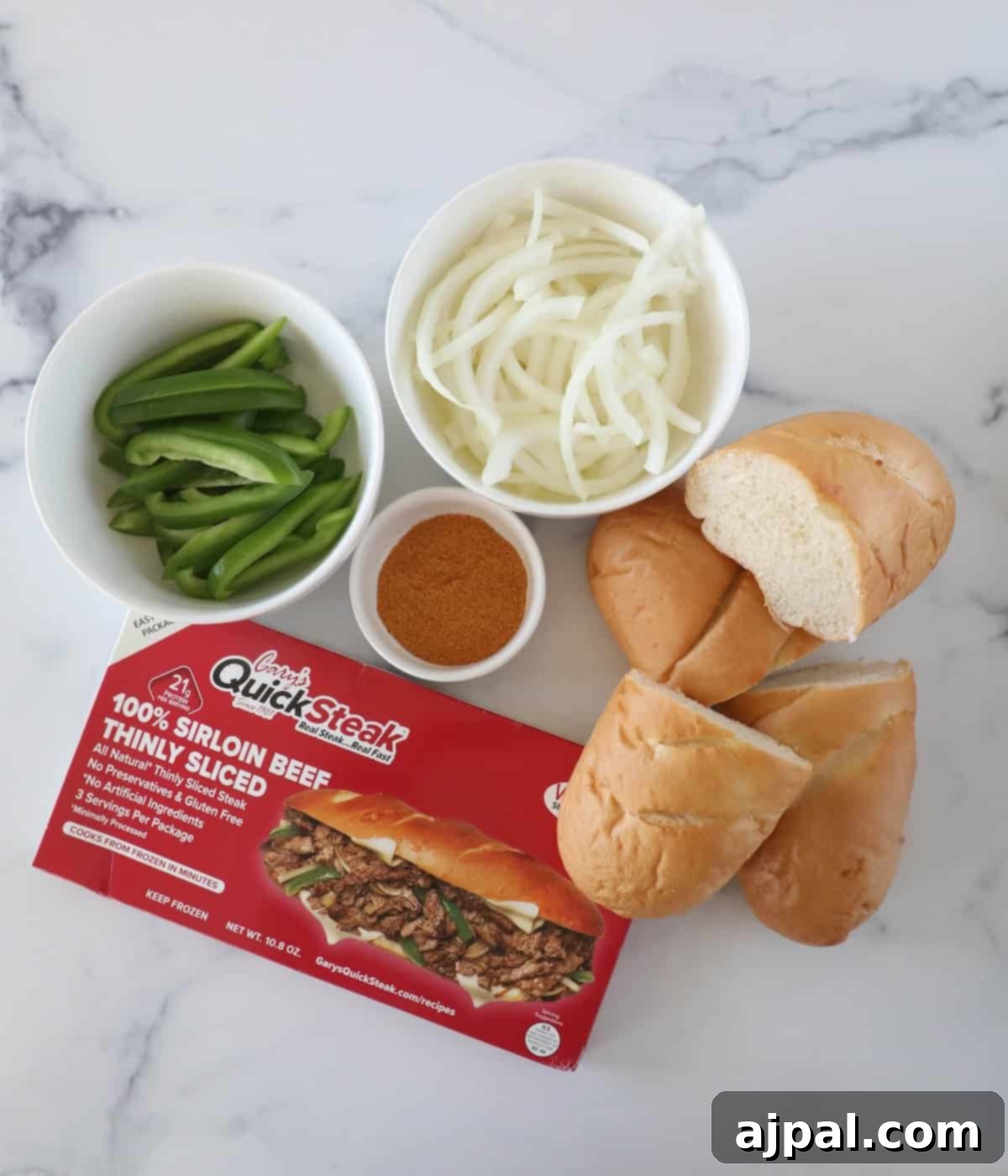 Ingredients for a sandwich: sliced green peppers, onions, seasoning, steak strips package, and hoagie rolls on a marble surface.