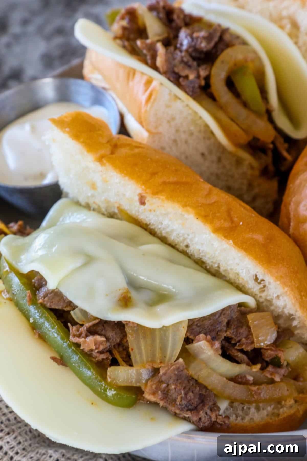 Close-up of a Philly cheesesteak sandwich with melted cheese, sautéed onions, and green peppers inside a hoagie roll. A small dish of sauce is on the side.