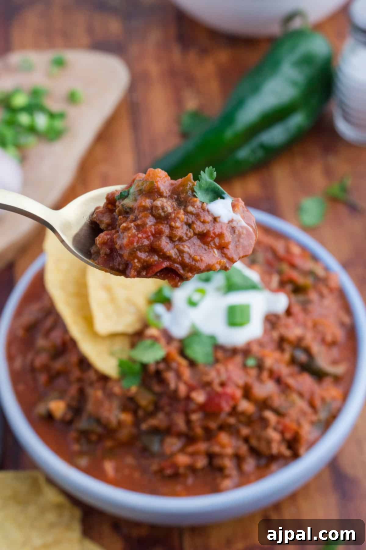 A spoonful of chili with cilantro and sour cream tops a bowl of hearty chili, garnished with crispy chips and vibrant green onions. A poblano pepper and cutting board add an authentic touch in the background.