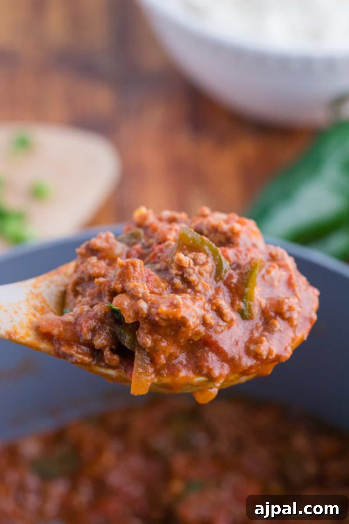 Close-up of a spoon lifting a portion of rich, red poblano chili with meat and poblano peppers in a rich tomato sauce, held over a pot.