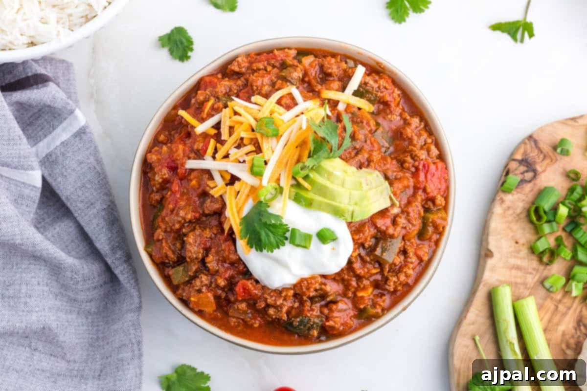 A bowl of chili brimming with bold flavors is topped with shredded cheese, creamy avocado slices, sour cream, cilantro, and green onions. Poblano peppers add a smoky twist to this comforting dish set alongside chopped green onions and a gray napkin.