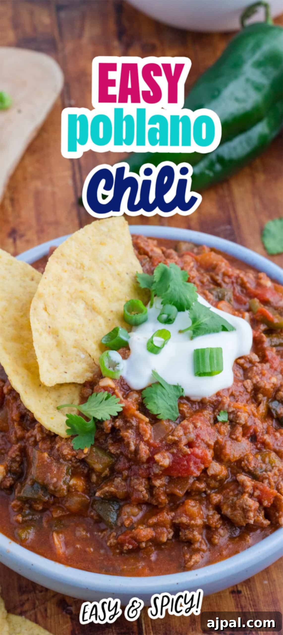 A bowl of savory chili with ground meat and beans, topped with sour cream, chopped green onions, fresh cilantro, and crunchy tortilla chips. In the background, a jalapeño hints at the rich flavors of this easy Poblano Chili.
