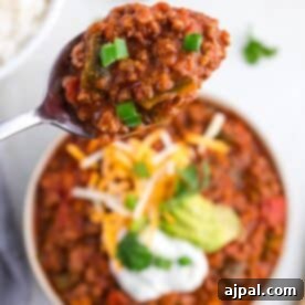A spoonful of chili with green onions held above a bowl of hearty poblano chili, topped with avocado slices, shredded cheese, and sour cream.