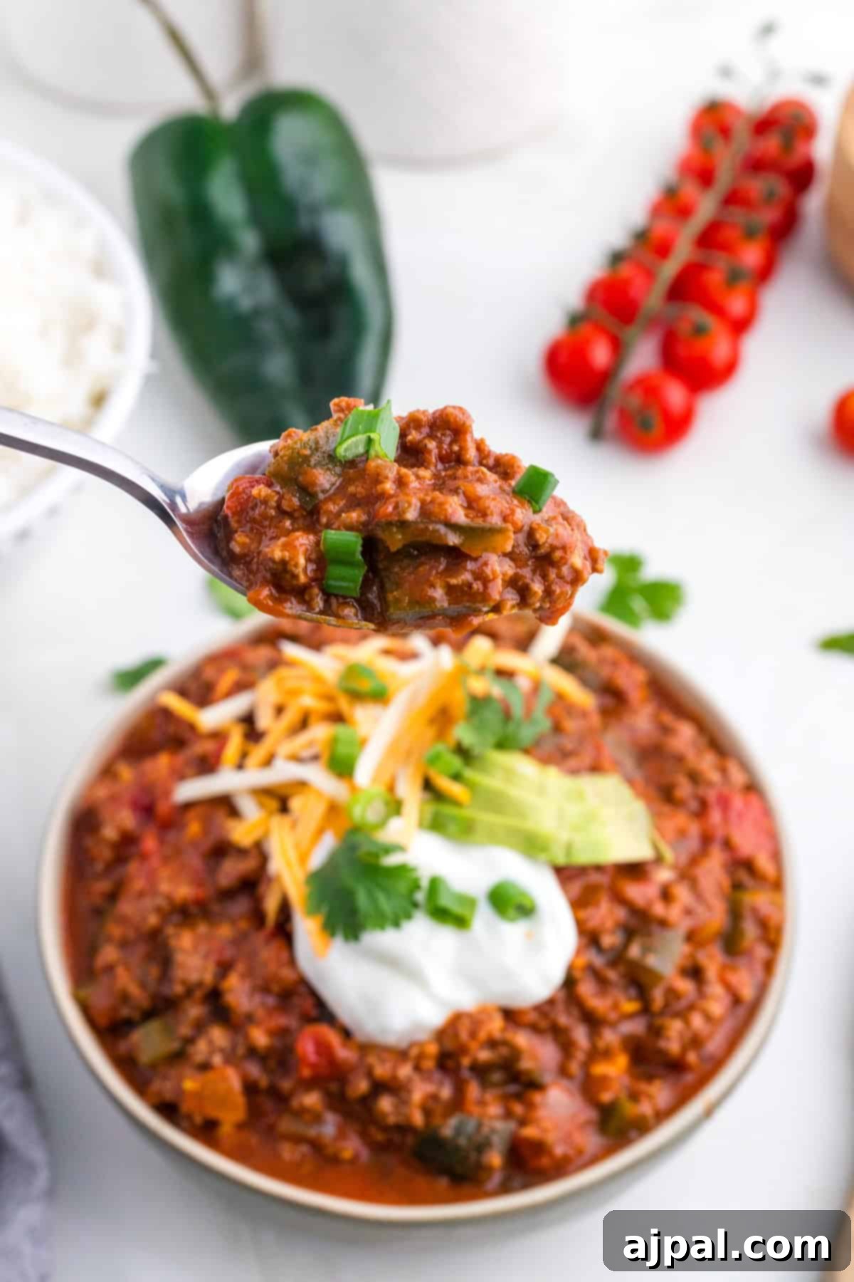 A spoonful of chili with ground meat and vegetables, accented by a spicy poblano kick, is held above a bowl. The dish is garnished with sour cream, shredded cheese, avocado slices, and green onions. Cherry tomatoes and a pepper are artfully arranged in the background.
