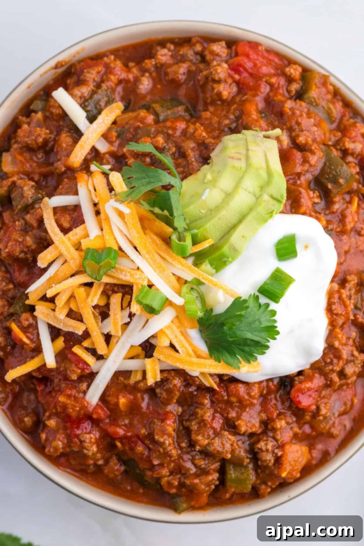 A hearty bowl of chili featuring smoky poblano peppers, topped with shredded cheese, creamy avocado slices, sour cream, and garnished with cilantro and green onions.