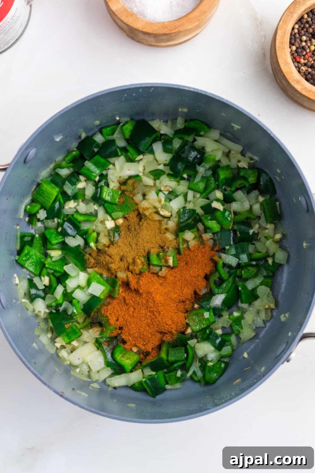 A pan with chopped poblano peppers and onions, sprinkled with ground spices, rests on a light countertop. Nearby, bowls of chili flakes and salt complete the flavorful setup.