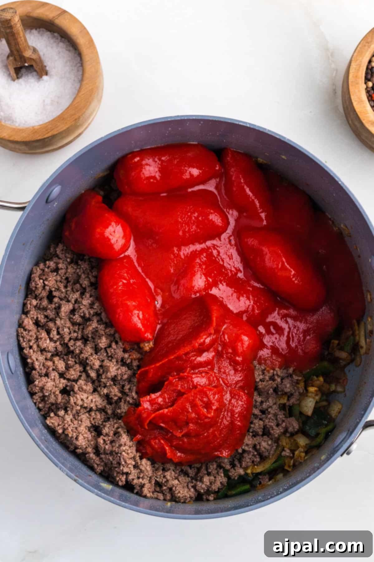 Ground beef, whole tomatoes, and a hint of poblano chili simmer in a pot, shown from above. Salt and pepper bowls are visible nearby.