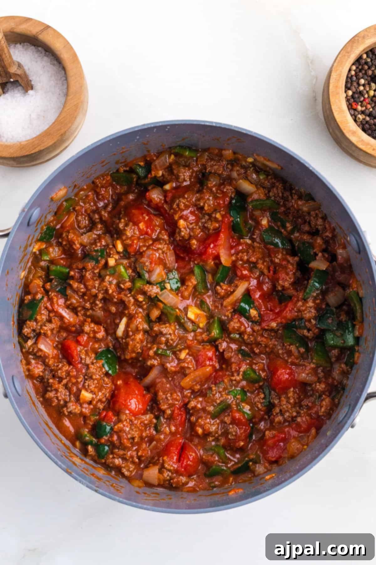 A pot of chili featuring ground meat, diced tomatoes, and chopped green peppers is seasoned to perfection. Beside it are two small wooden bowls with salt and pepper. A hint of poblano chili adds a subtle smoky flavor, elevating the dish to new heights.