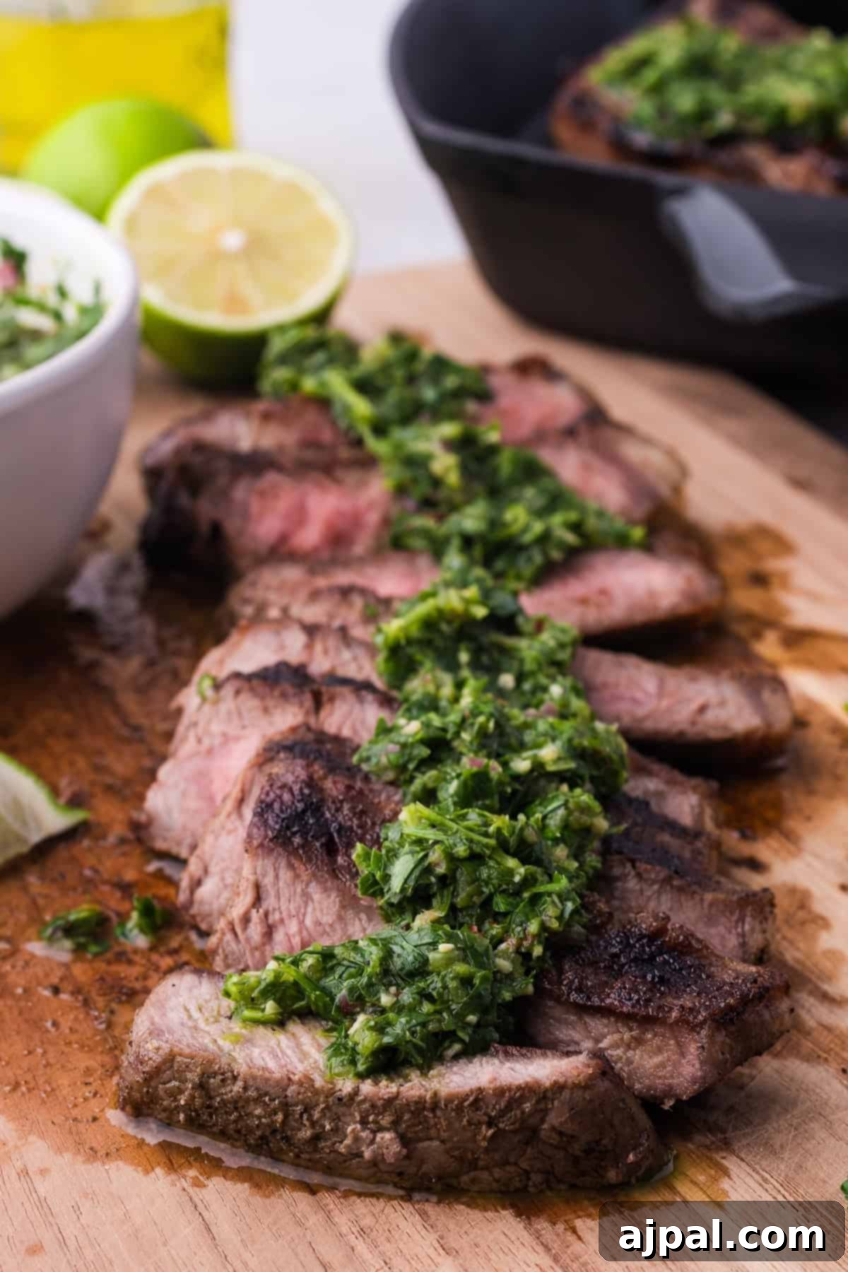 Steak with Chimichurri 2 Grilled steak slices topped with chimichurri sauce rest on a wooden board, paired with a halved lime and a bowl of green garnish.