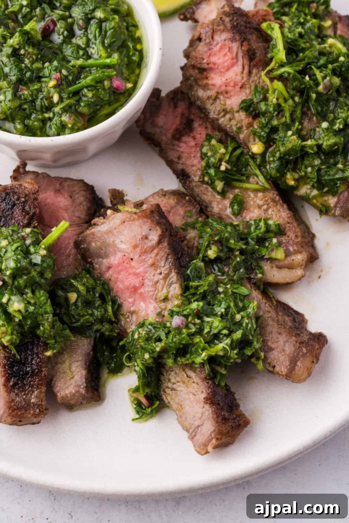 Steak with Chimichurri 3 Sliced grilled steak draped in zesty green chimichurri sauce on a pristine white plate, accompanied by a small bowl of extra chimichurri for those craving more of its vibrant flavor. Perfectly crafted for this mouthwatering steak recipe.