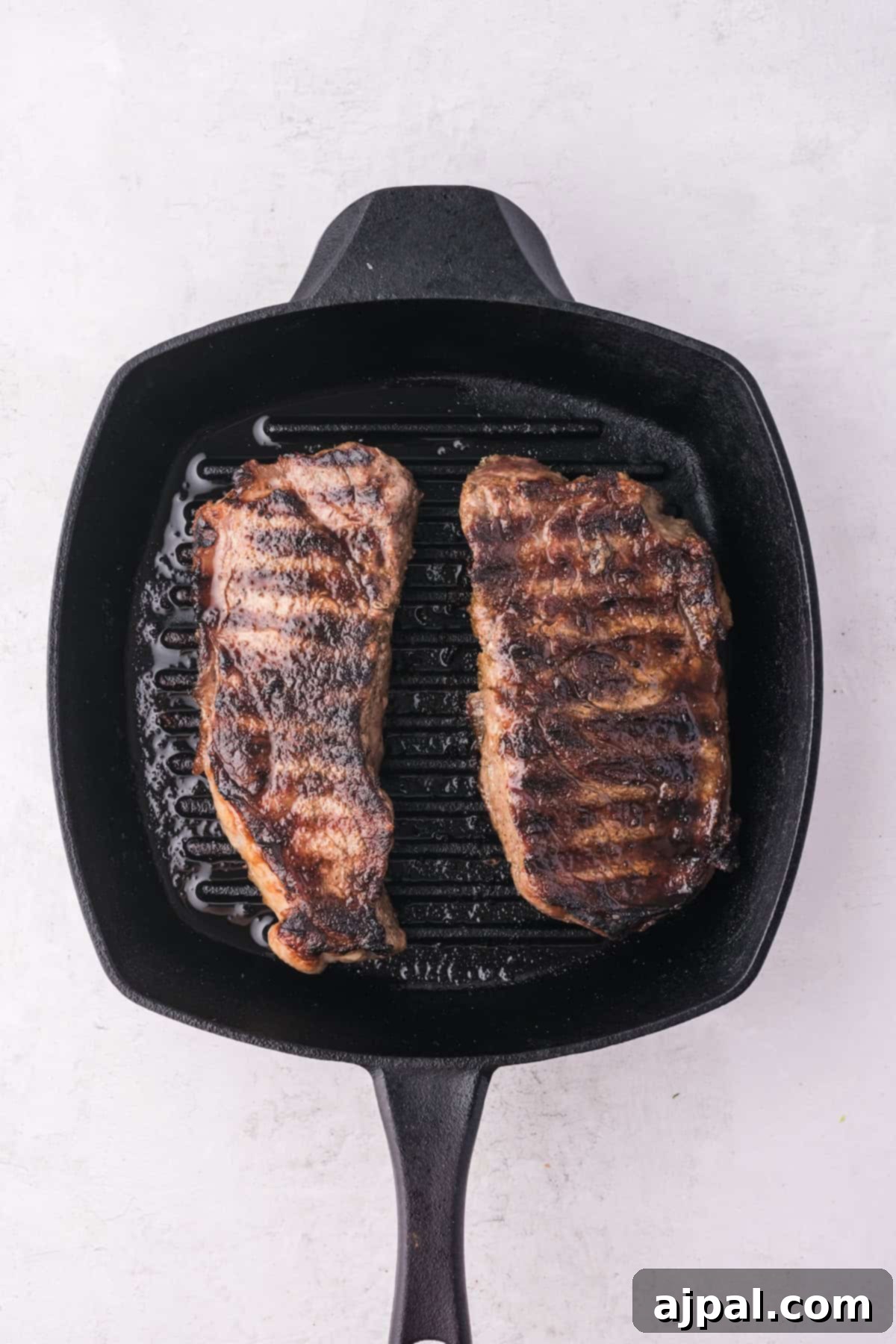 Steak with Chimichurri 8 Two grilled steak pieces with sear marks sizzle in a black square grill pan on a light background, perfectly paired with vibrant chimichurri.