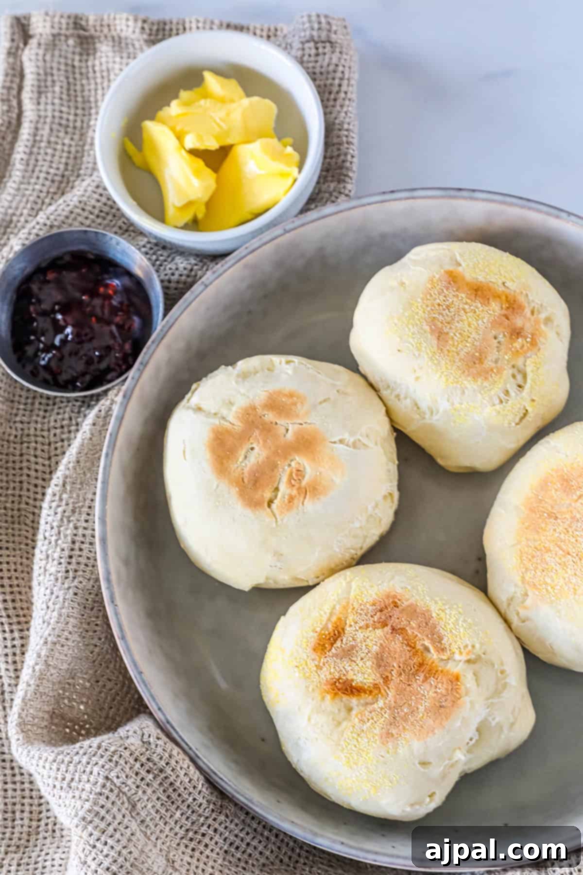 Four English muffins on a plate with dishes of butter and jam nearby, placed on a burlap cloth.