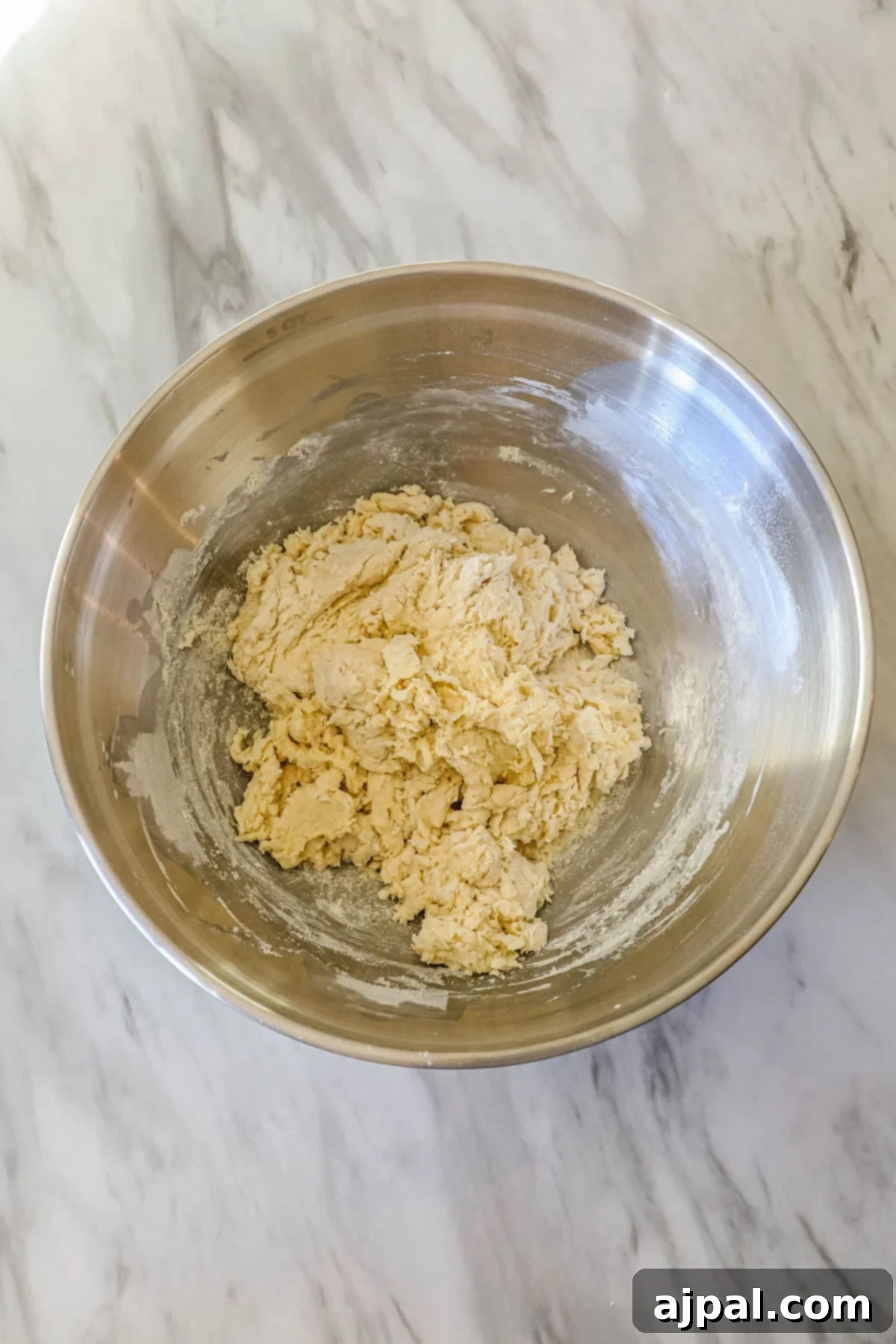 Dough mixture in a stainless steel bowl placed on a marbled countertop.