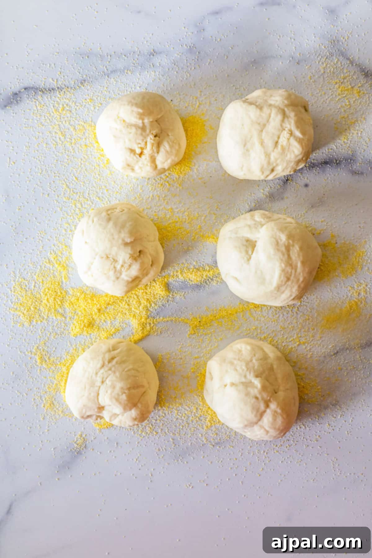 Six balls of dough are arranged on a floured marble surface, each dusted with yellow cornmeal.