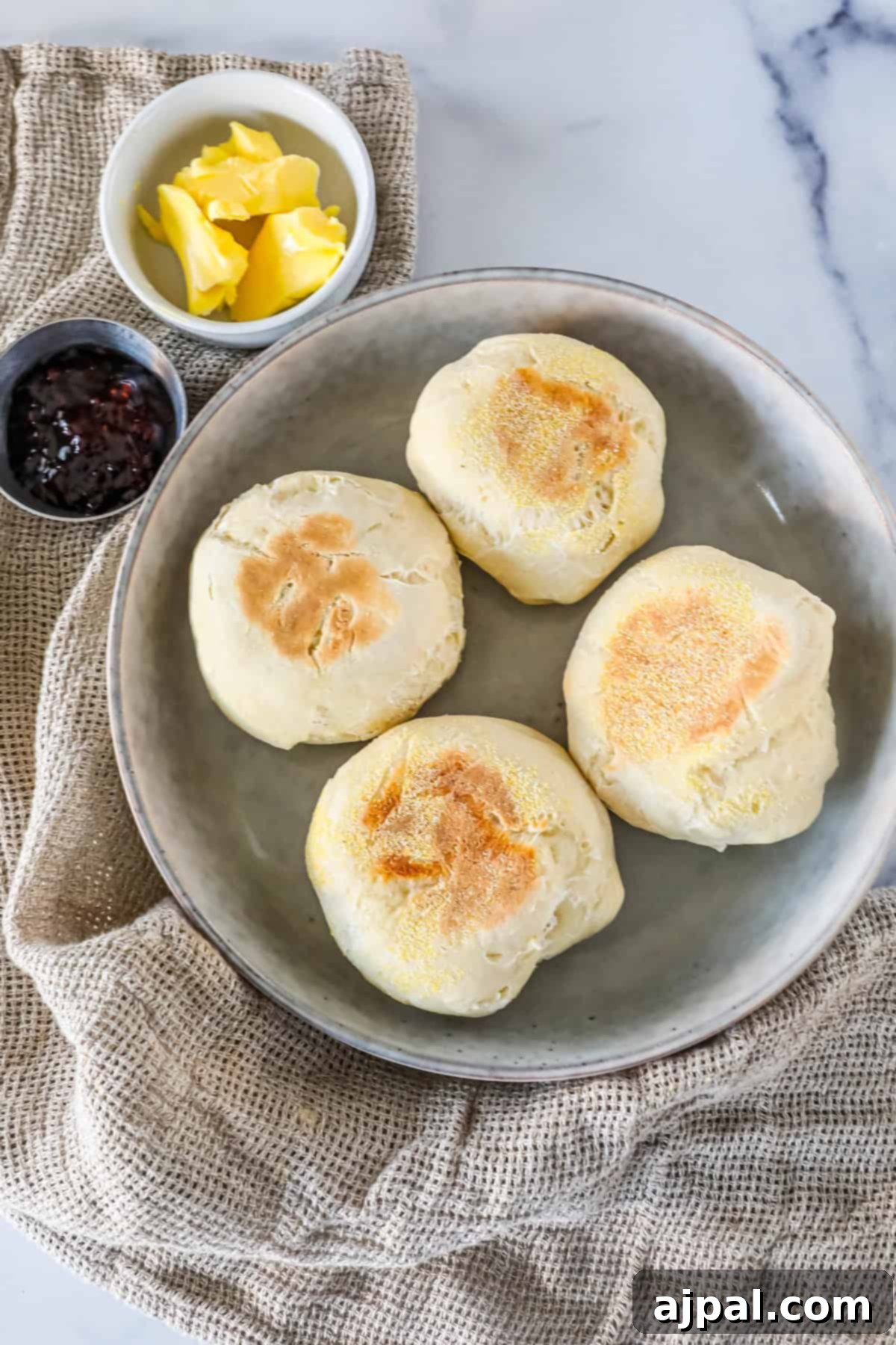 Four English muffins on a plate with a small bowl of butter and a small cup of jam on a textured cloth background.