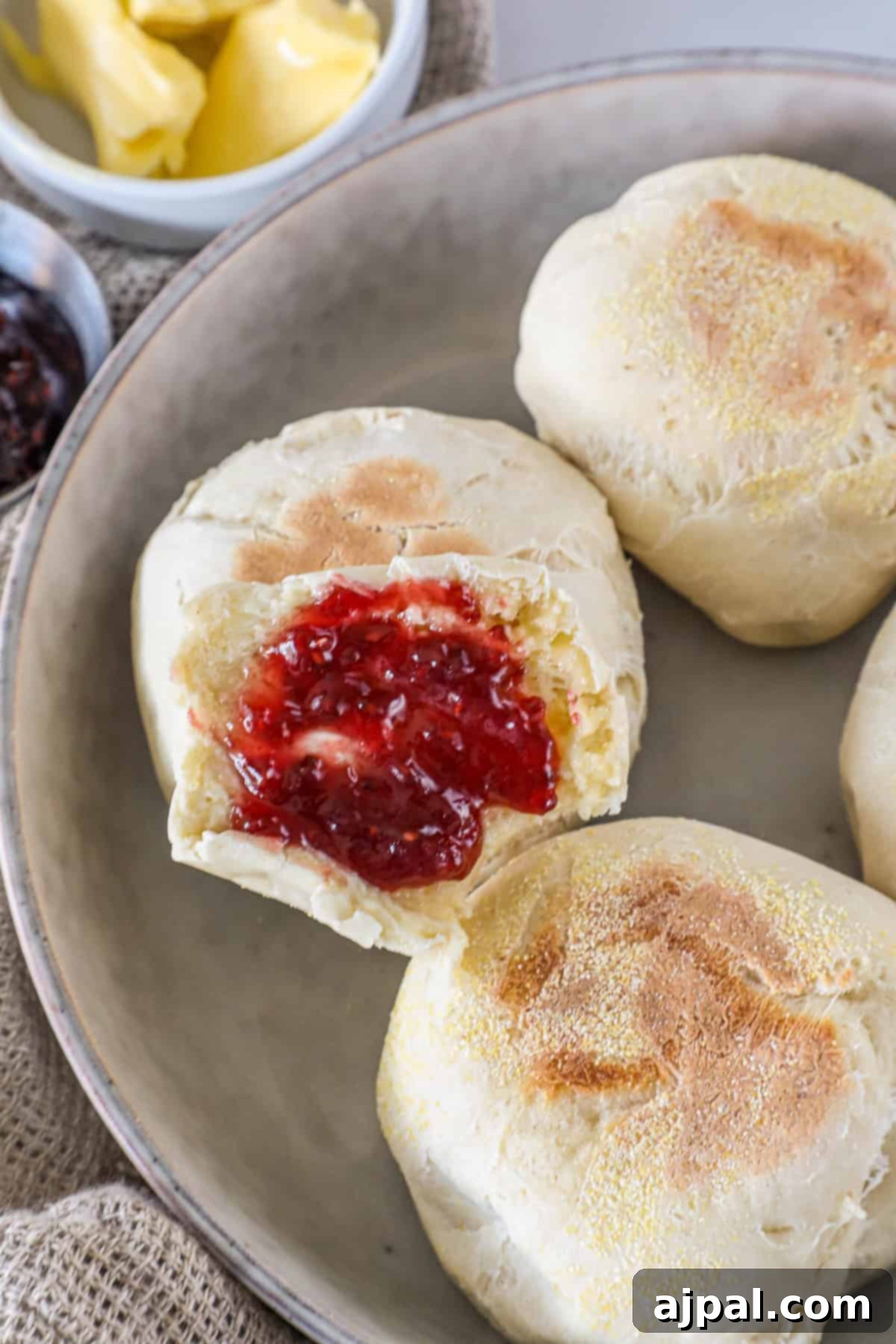 A plate with English muffins, one topped with red jam. Butter is in a small dish nearby.