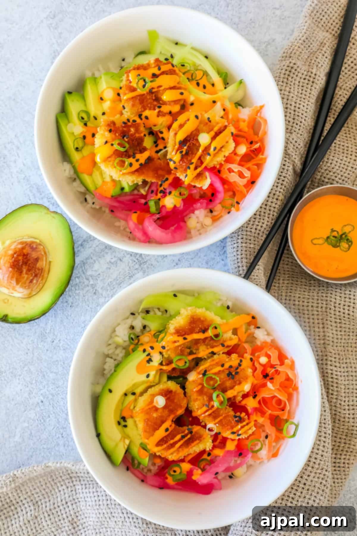 Golden Crispy Shrimp Rice Bowls 11 Two white bowls filled with fluffy rice, topped with sliced avocado, sweet mango, golden crispy shrimp, tangy pink pickled onions, fresh scallions, and toasted sesame seeds. A creamy orange sauce in a cup and a sliced avocado are placed next to the bowls.