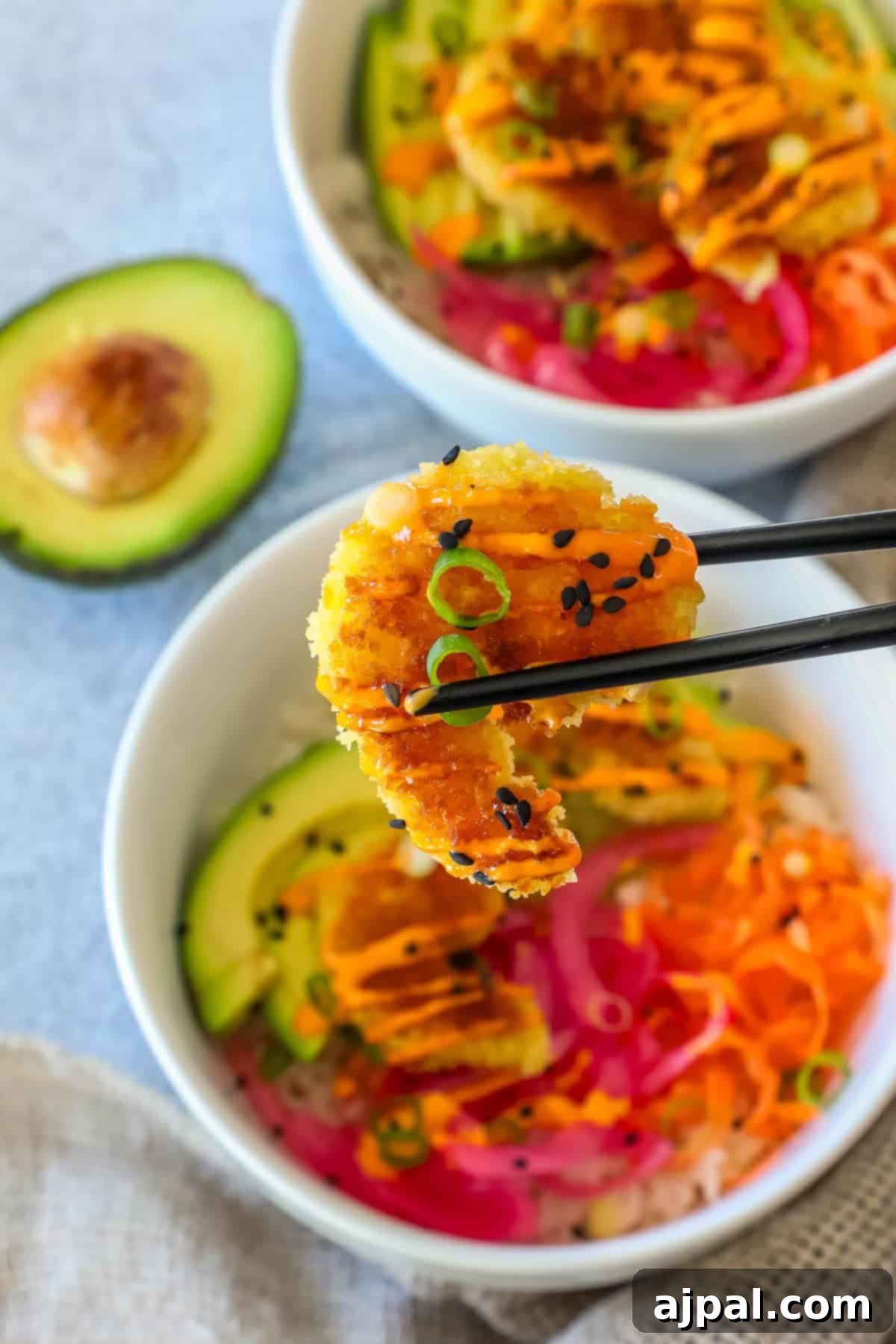 Golden Crispy Shrimp Rice Bowls 3 Chopsticks delicately holding a piece of perfectly breaded crispy shrimp over a vibrant rice bowl, showcasing layers of sliced avocado, bright pickled onions, and a rich, drizzled sauce. A fresh avocado half rests in the soft-focus background.