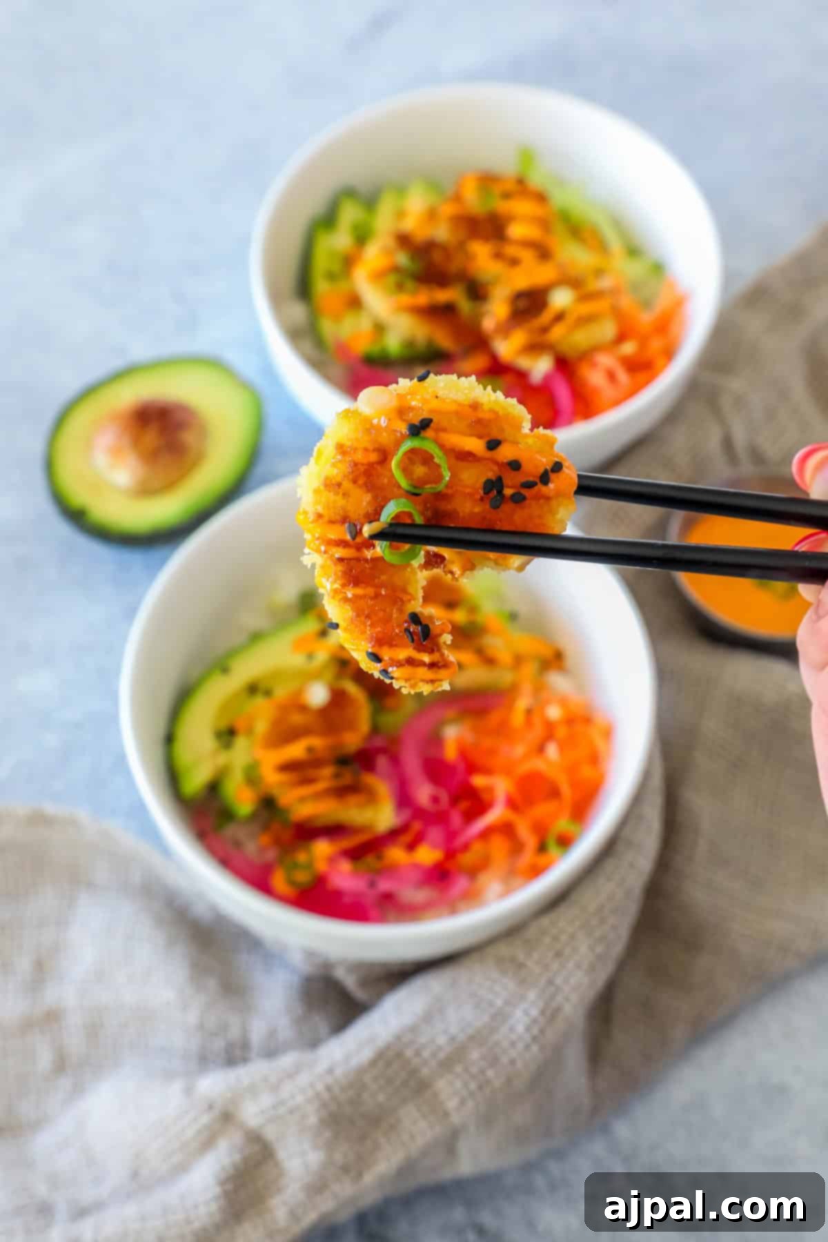 Golden Crispy Shrimp Rice Bowls 9 Chopsticks holding a single breaded shrimp, poised above two inviting bowls of fresh salad, each featuring creamy avocado, vibrant carrots, and a delicious sauce. A half avocado and a small container of dressing are visible nearby, highlighting the fresh ingredients.