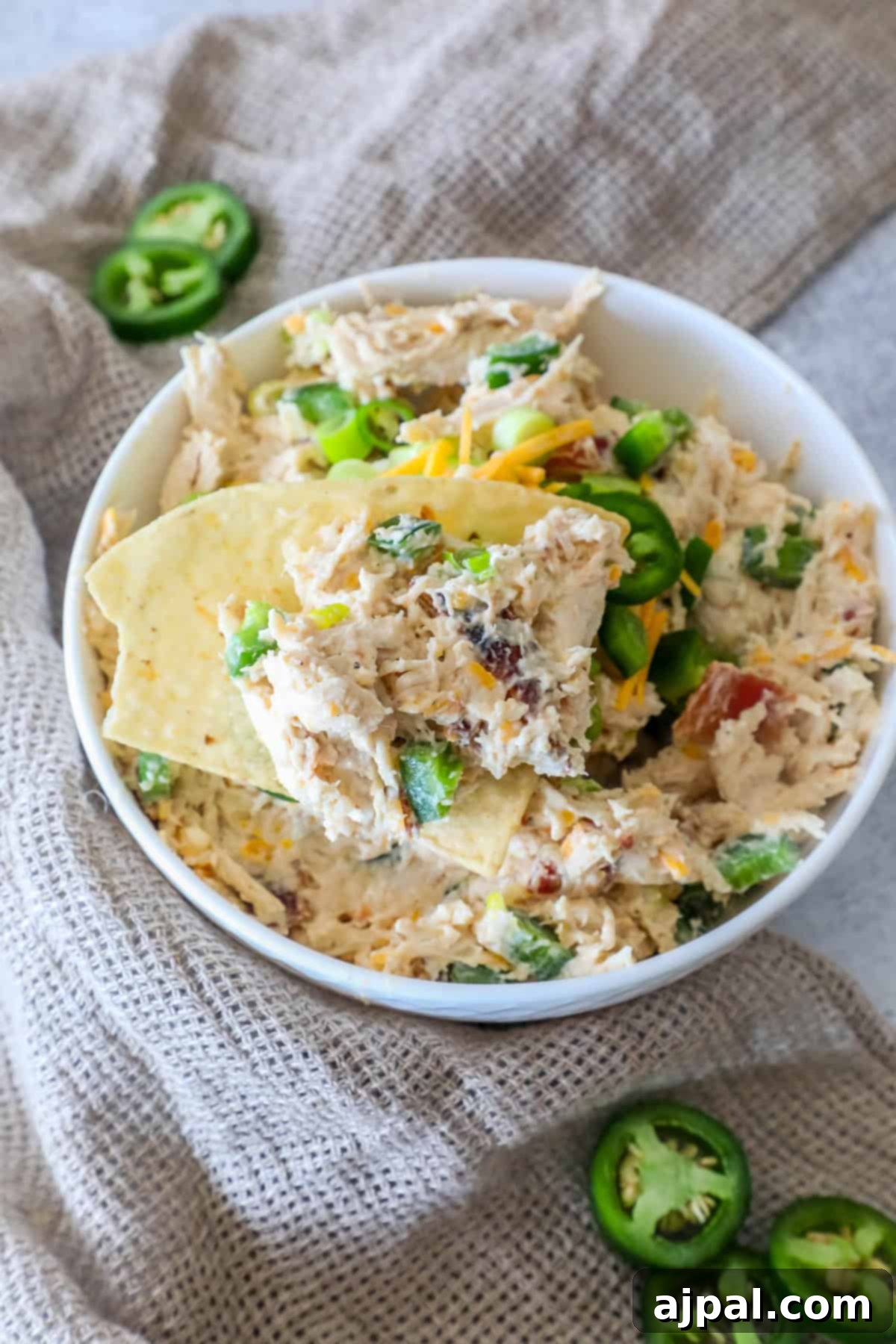 Ultimate Chicken Jalapeno Popper Dip 2 Bowl of creamy chicken dip with sliced jalapeños and shredded cheese, garnished with tortilla chips, set on a textured cloth. This is the ultimate chicken jalapeno popper dip for any gathering.