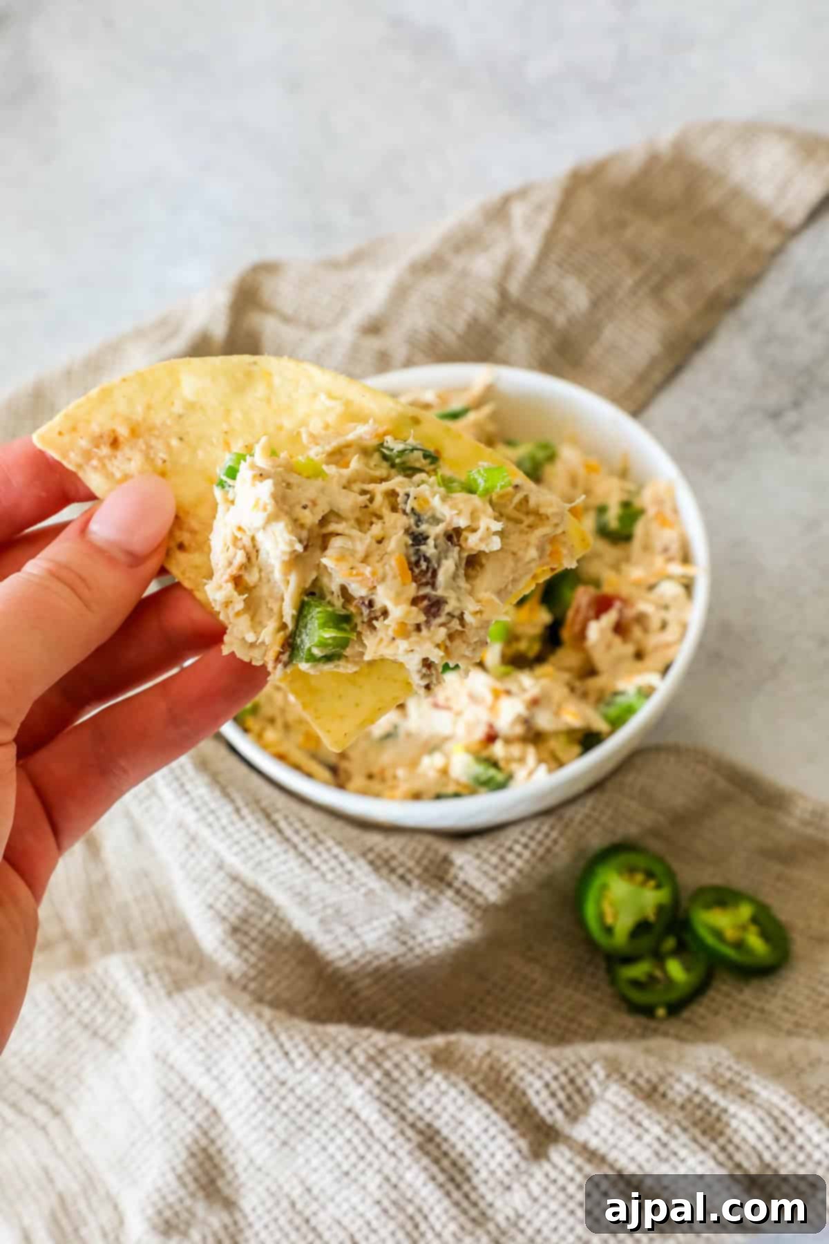 Creamy Chicken Jalapeno Popper Dip 10 A hand holds a tortilla chip with creamy dip, topped with vegetables, over a dish. Sliced jalapeños are on the side.
