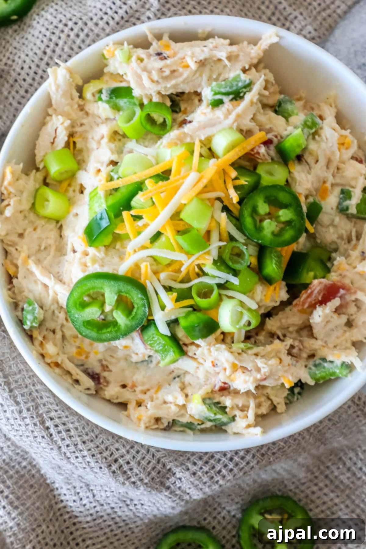 Ultimate Chicken Jalapeno Popper Dip 3 A bowl of chicken dip topped with sliced jalapeños, green onions, and shredded cheese, placed on a burlap surface. This creamy dip is perfect for any occasion.