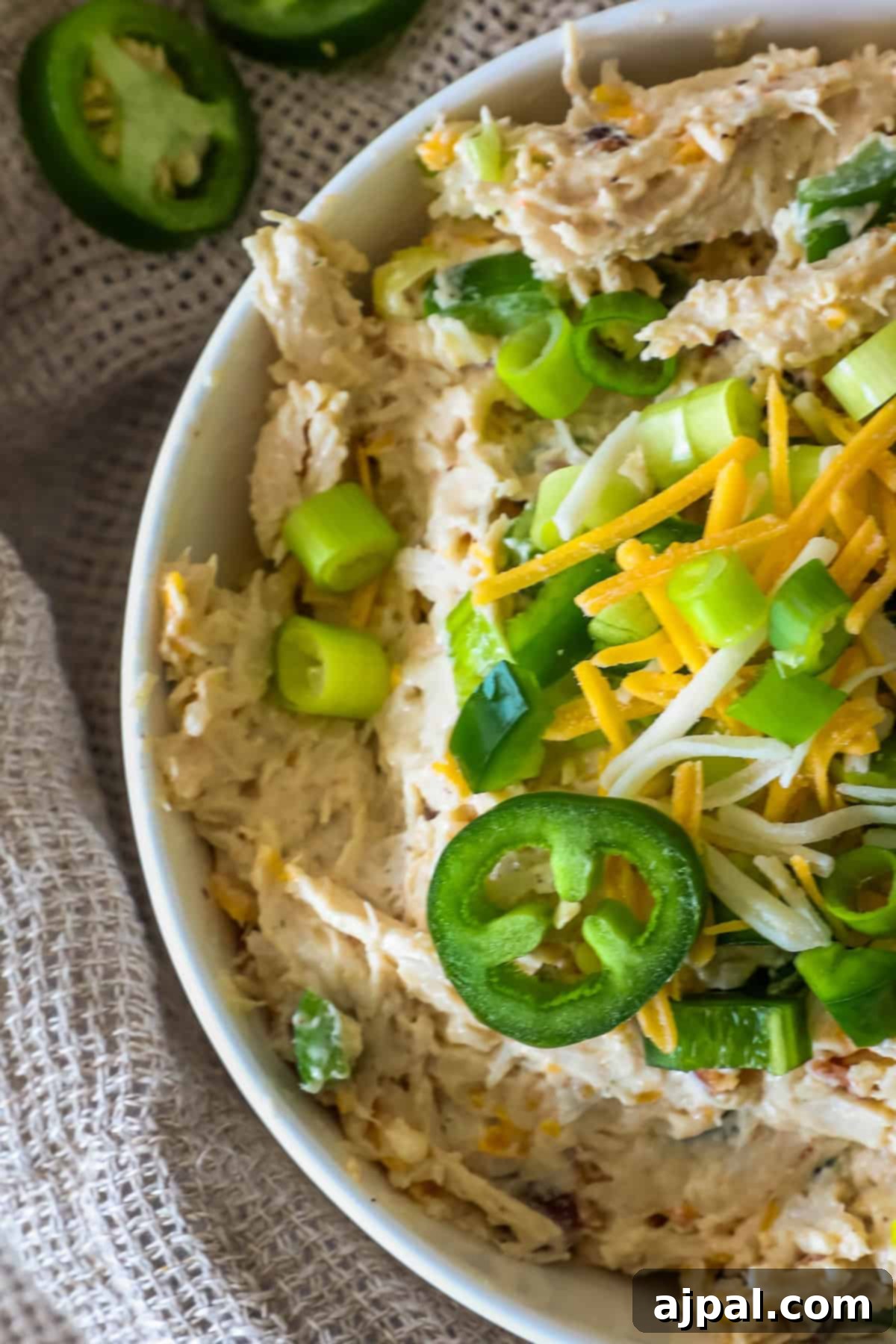 Creamy Chicken Jalapeno Popper Dip 4 A bowl of creamy dip topped with sliced jalapeños, shredded cheese, and chopped green onions on a textured cloth background.