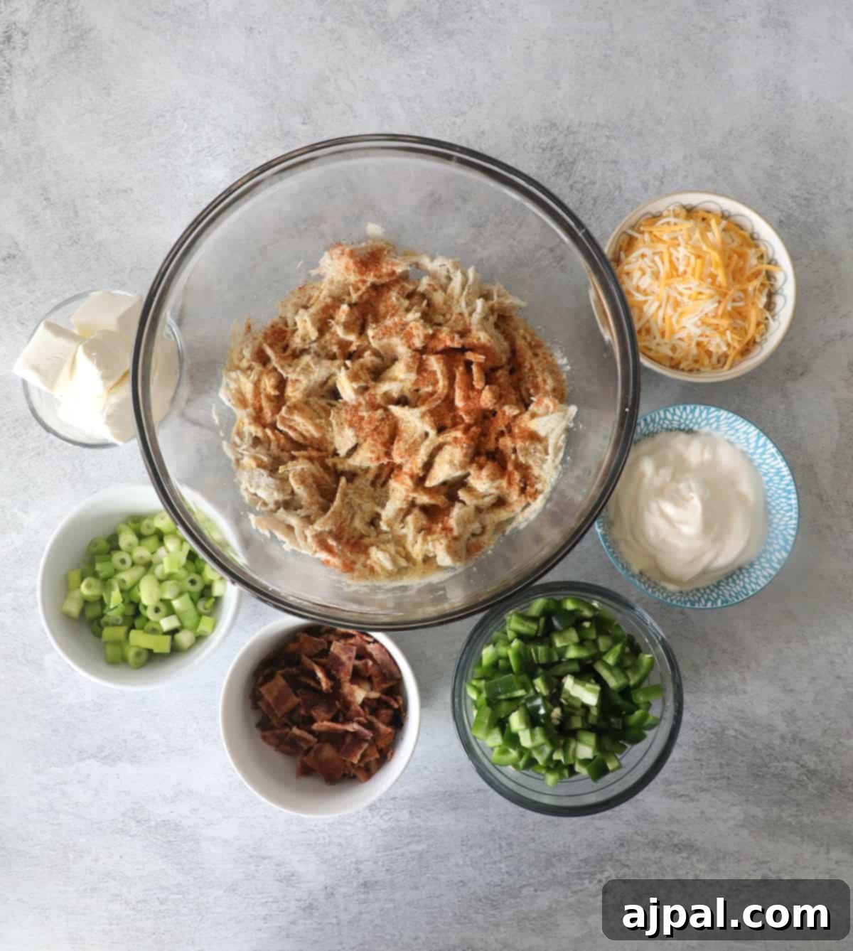 Creamy Chicken Jalapeno Popper Dip 5 Ingredients for a dip arranged on a table: a bowl of shredded chicken with spices, and bowls of cream cheese, shredded cheese, sour cream, green onions, bacon, and diced green peppers.