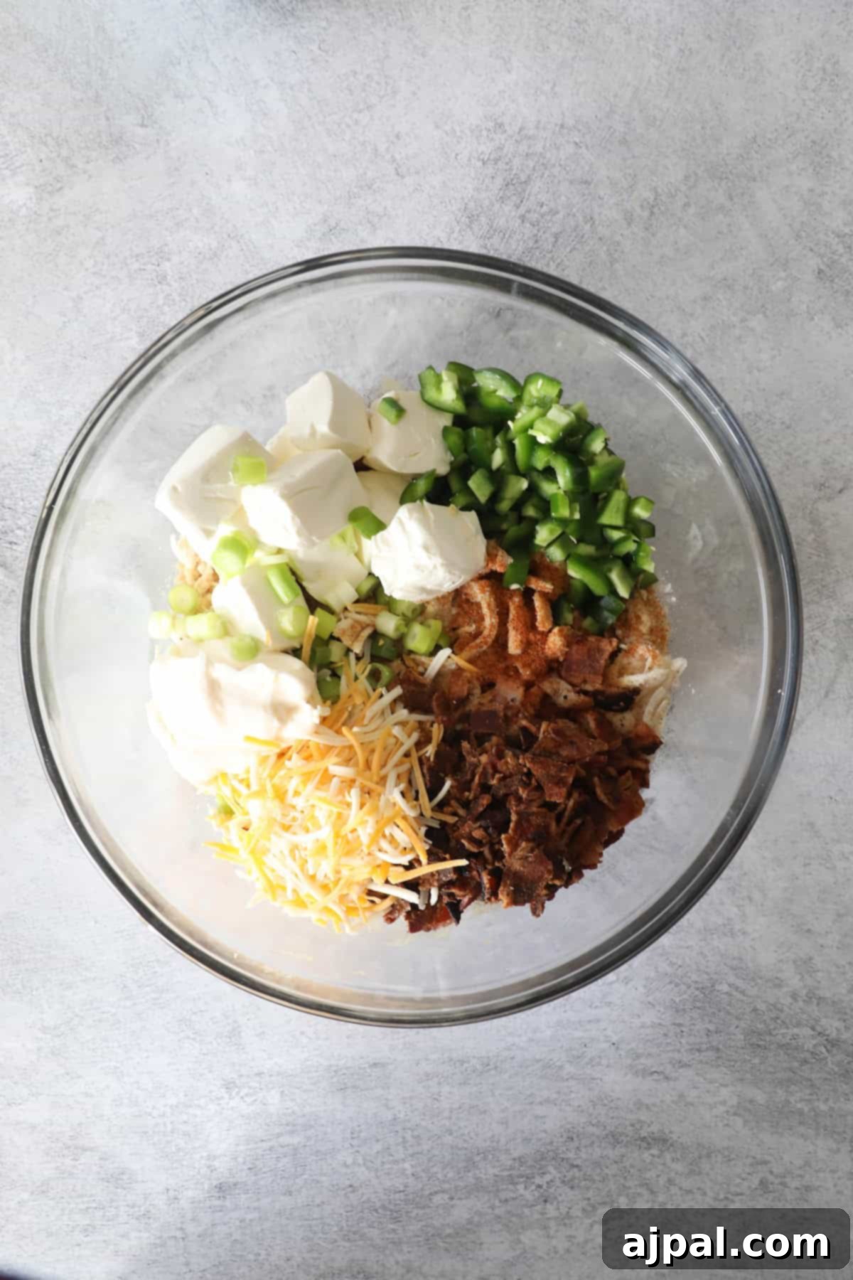 Creamy Chicken Jalapeno Popper Dip 6 A glass bowl containing cream cheese, diced green peppers, shredded cheese, chopped bacon, and sour cream, placed on a light gray surface.