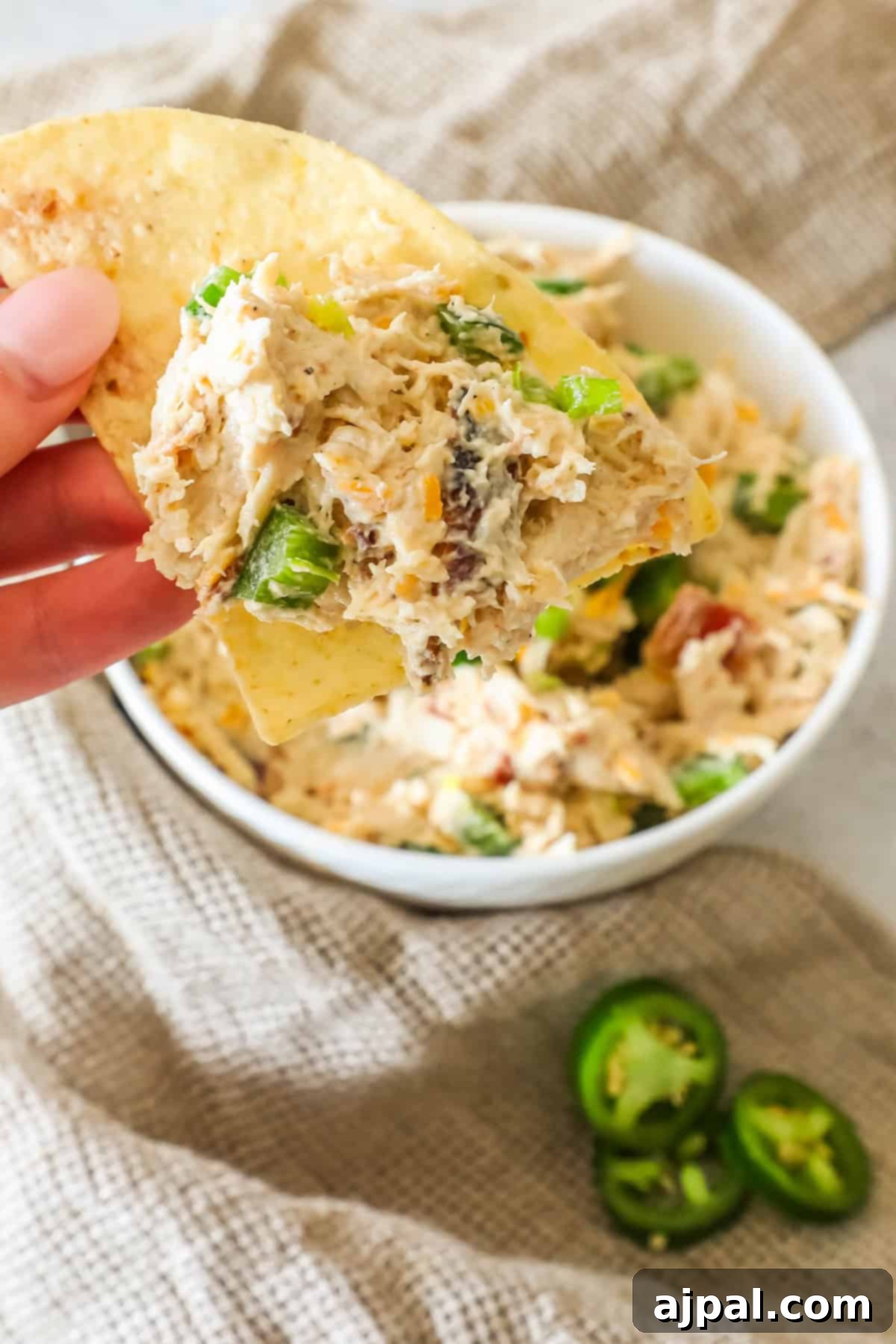 Creamy Chicken Jalapeno Popper Dip 8 A hand holding a tortilla chip topped with creamy dip over a bowl. Sliced jalapeños and a cloth are in the background.