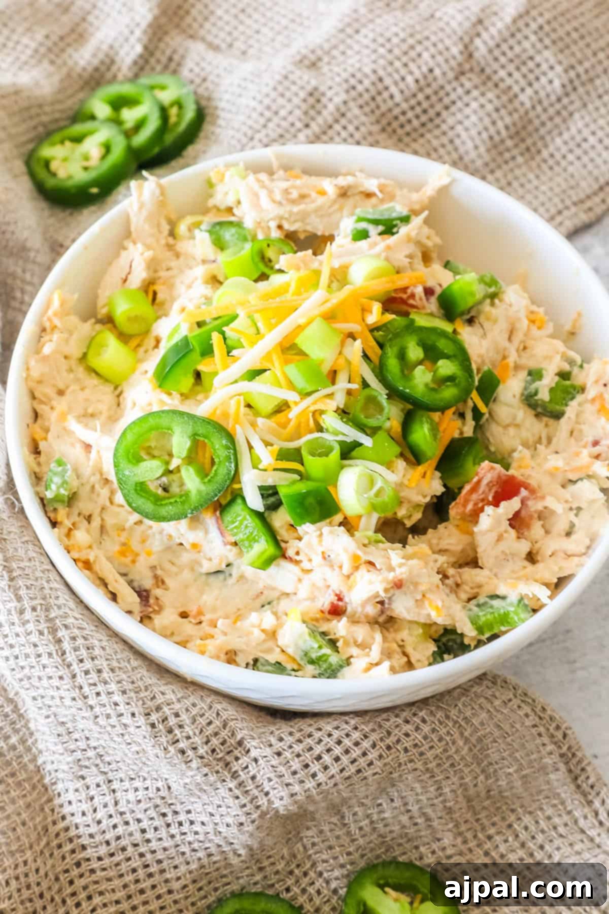 Creamy Chicken Jalapeno Popper Dip 9 A bowl of chicken salad topped with sliced jalapeños, shredded cheese, and chopped green onions on a burlap cloth.