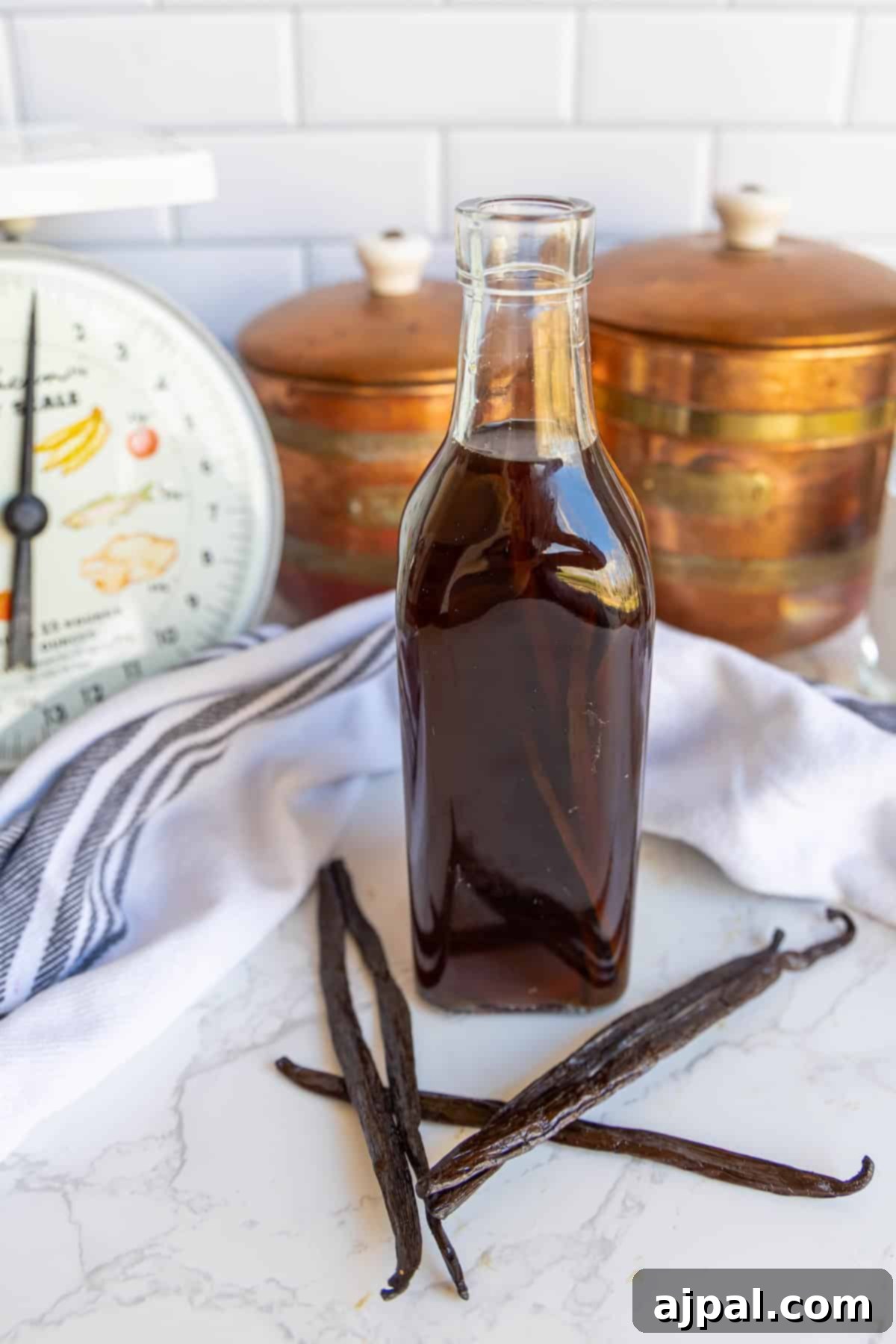 A bottle of dark liquid stands on a marble surface, surrounded by vanilla pods. A striped cloth, kitchen scale, and copper containers are in the background.