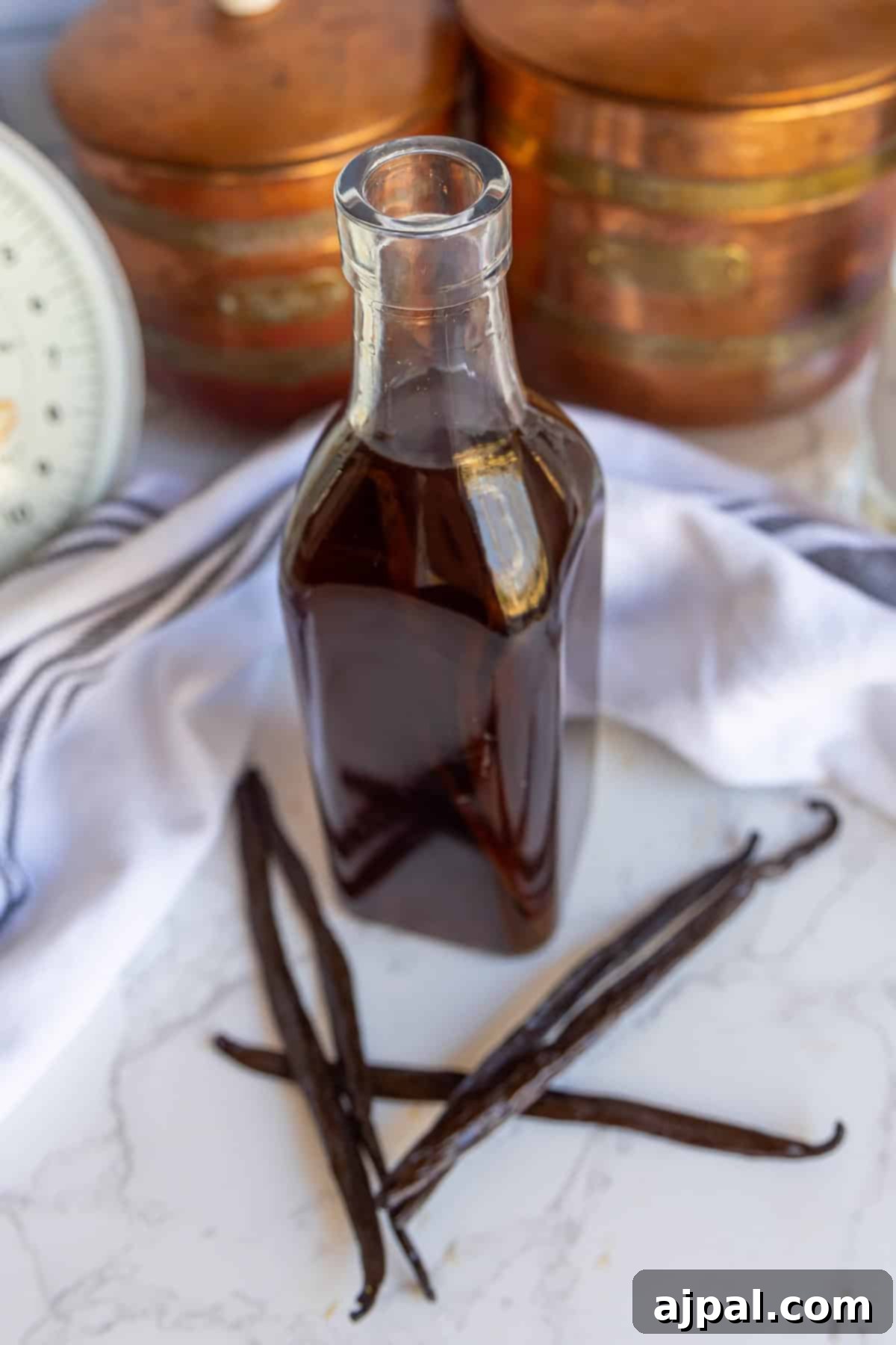 A glass bottle of vanilla extract beside vanilla pods on a marble surface with a striped cloth and vintage containers in the background.