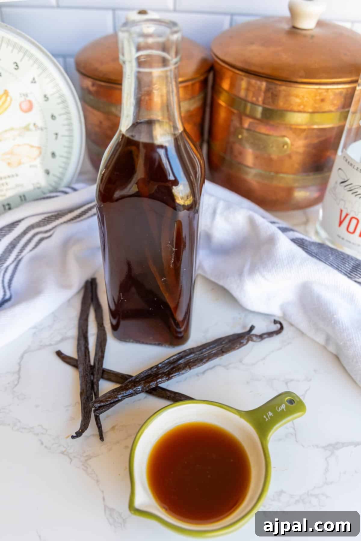 A glass bottle of dark liquid, vanilla beans, a small cup of similar liquid, copper canisters, a scale, and a vodka bottle on a white countertop.
