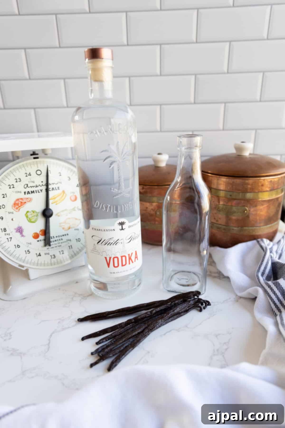 A bottle of vodka and an empty bottle on a kitchen counter, alongside dried vanilla beans, a vintage scale, two copper containers, and a striped cloth.