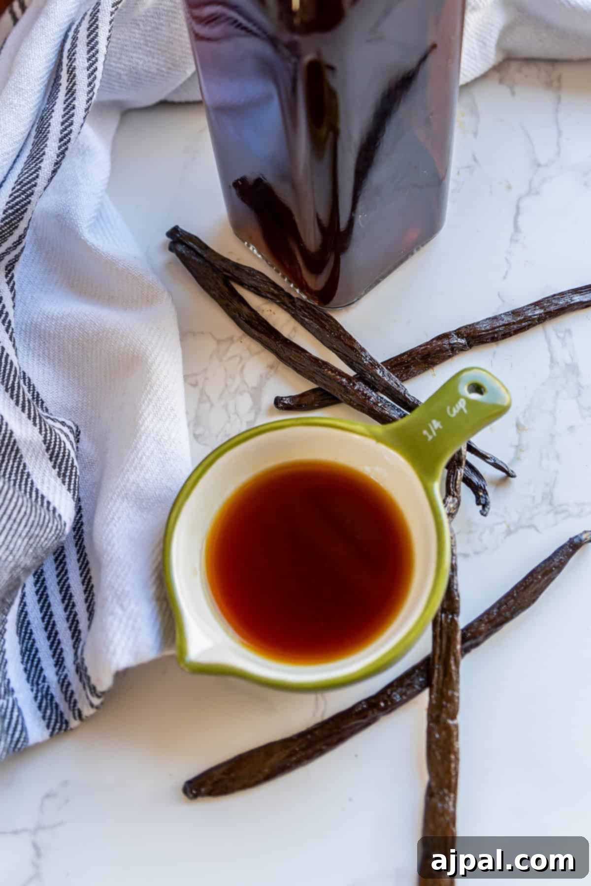 A small green measuring cup with vanilla extract is surrounded by vanilla beans and a striped cloth on a marble surface. A tall brown bottle is in the background.
