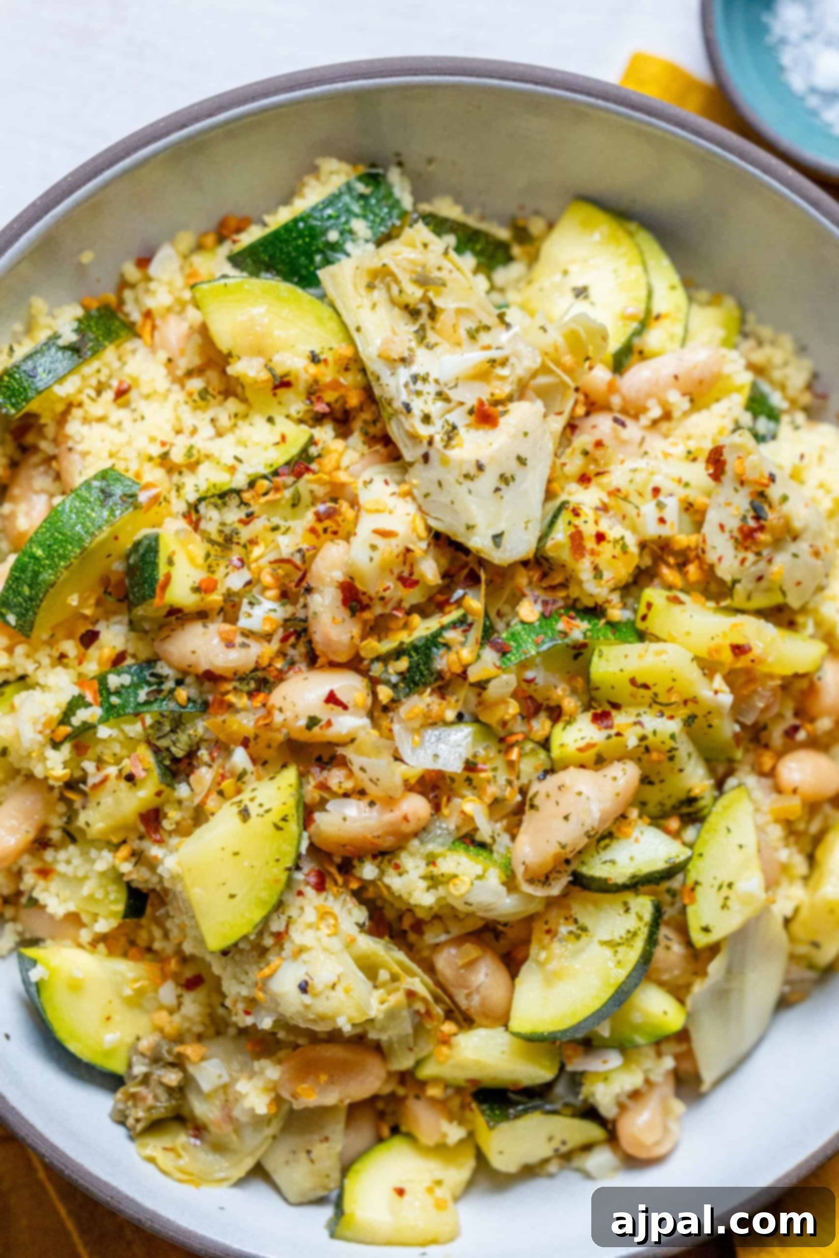 Spicy Mediterranean Cured Lemon Couscous 14 A beautifully presented bowl of spicy Mediterranean lemon couscous, featuring a vibrant mix of fluffy couscous, tender zucchini, white beans, and artichoke hearts. Garnished with fresh green herbs and a sprinkle of red pepper flakes, the dish looks inviting and perfectly balanced, highlighting its fresh ingredients and rich flavors.