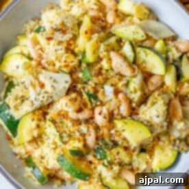 A bowl of couscous mixed with zucchini, white beans, artichoke hearts, and herbs, garnished with red pepper flakes.
