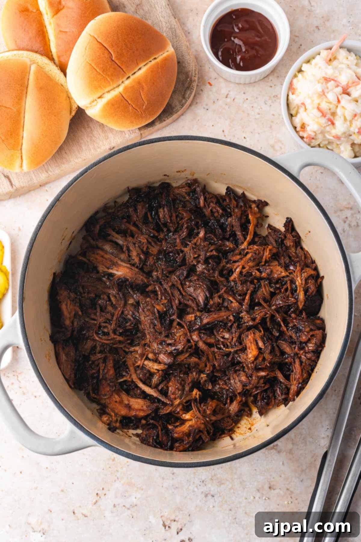 A Dutch oven brimming with tender pulled pork sits beside a wooden board adorned with buns, a dish of coleslaw, and a bowl of barbecue sauce, perfect for crafting that classic recipe everyone loves.