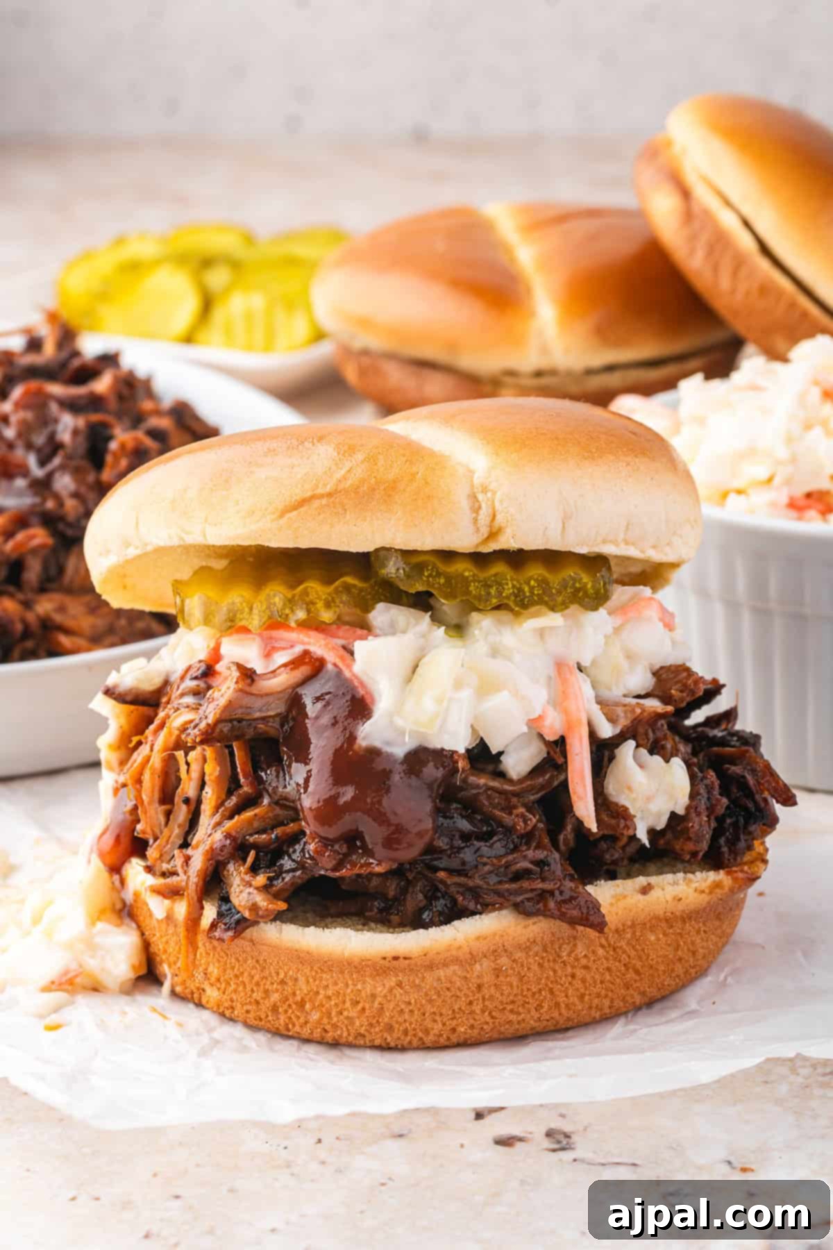Pulled pork sandwich crafted in a Dutch oven, topped with coleslaw, pickles, and barbecue sauce on a bun. Extras of buns, pickled slices, tender pulled pork, and coleslaw in the background.